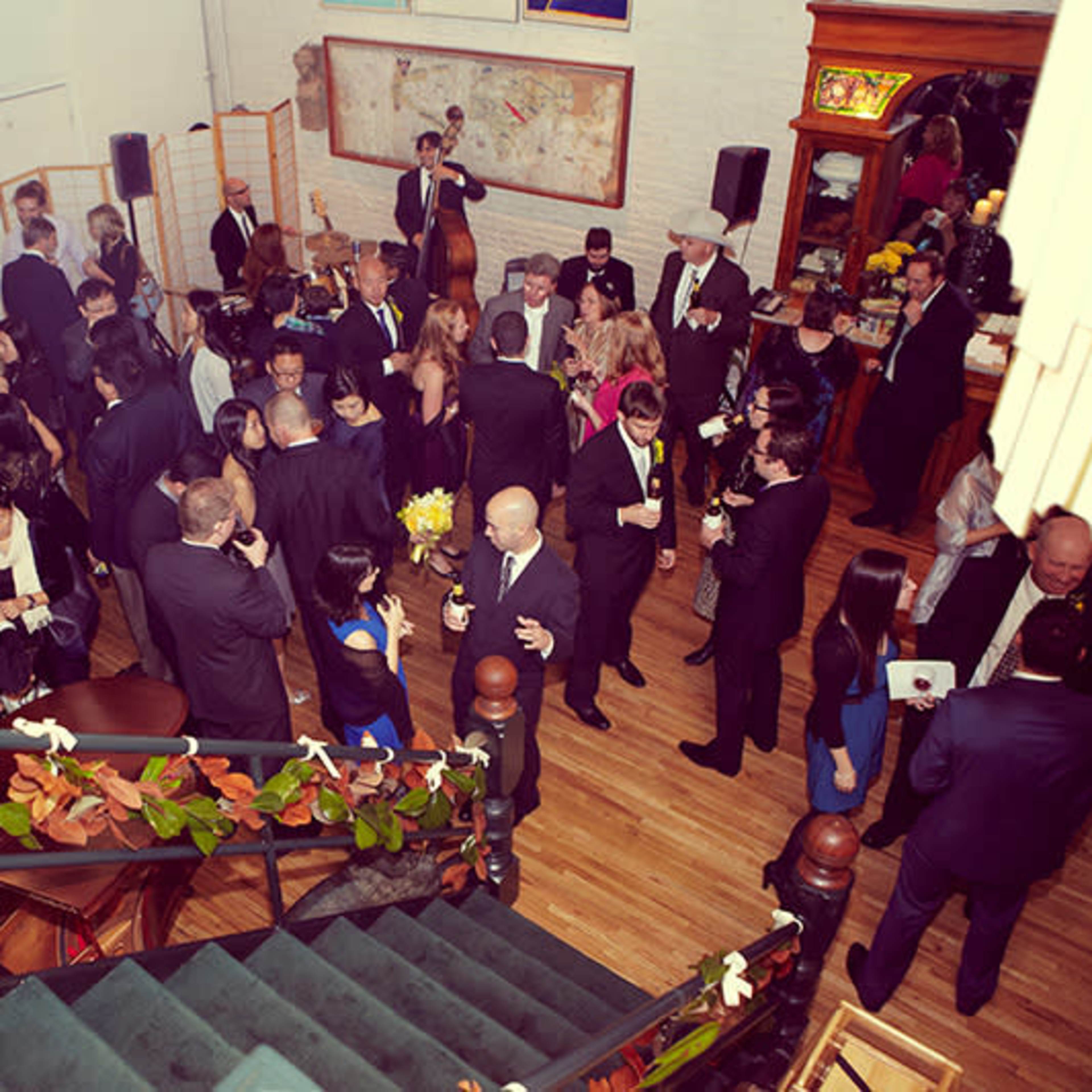 A large group of elegantly dressed guests mingles at a bustling event in a spacious indoor venue with art on the walls.