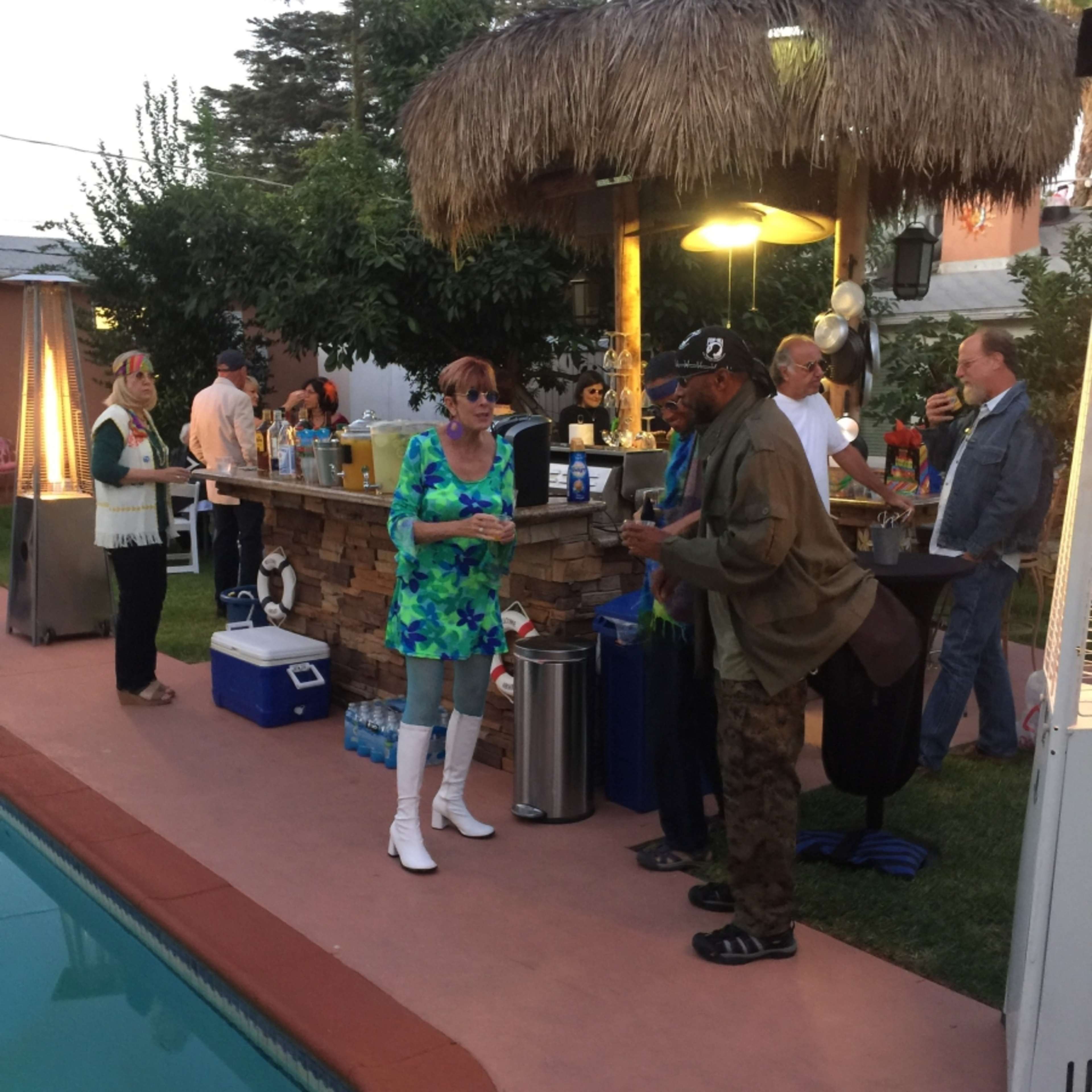 A group of people socializes around a poolside bar set under a thatched roof in a backyard.