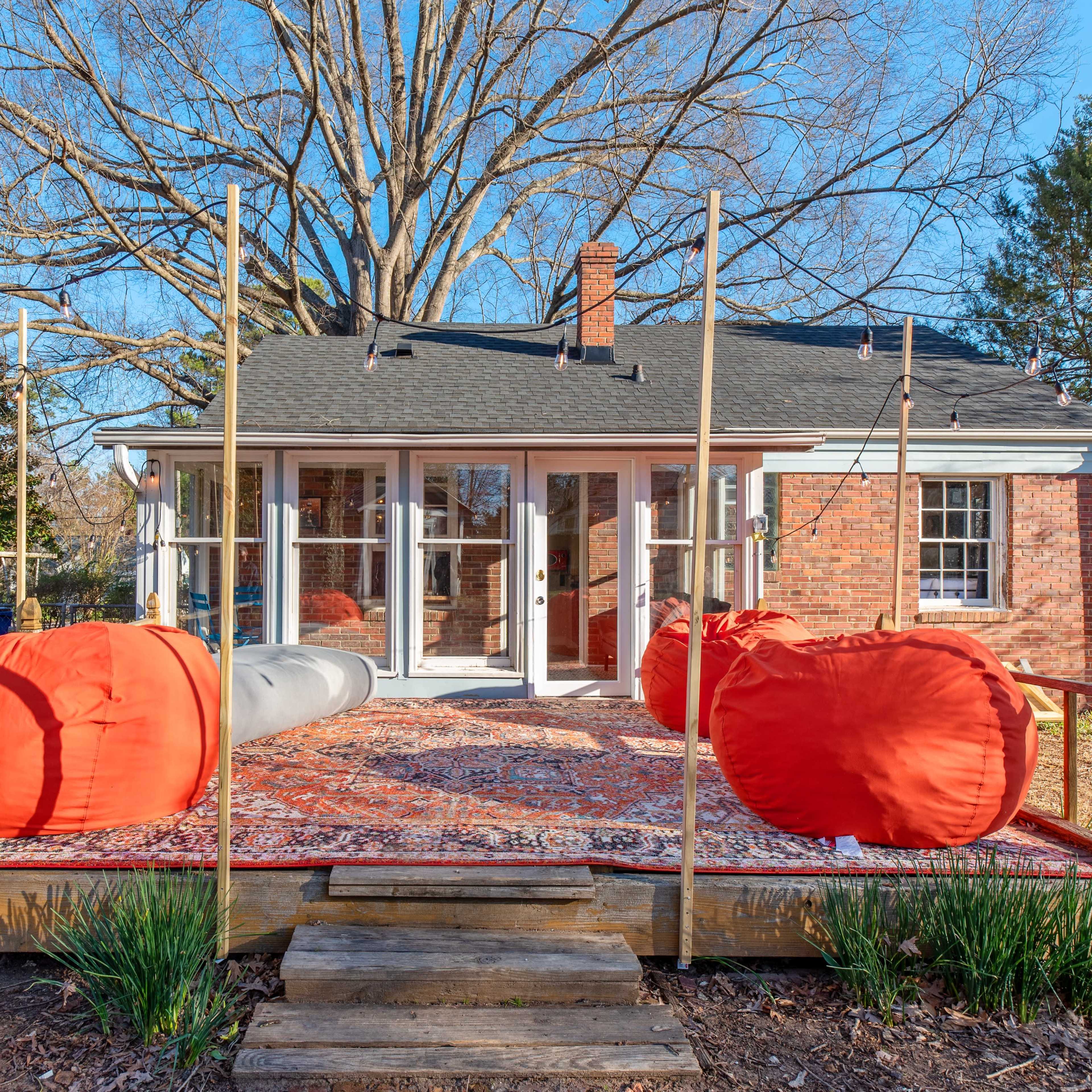A wooden deck with orange bean bag chairs and a patterned rug is positioned in front of a brick house surrounded by trees.