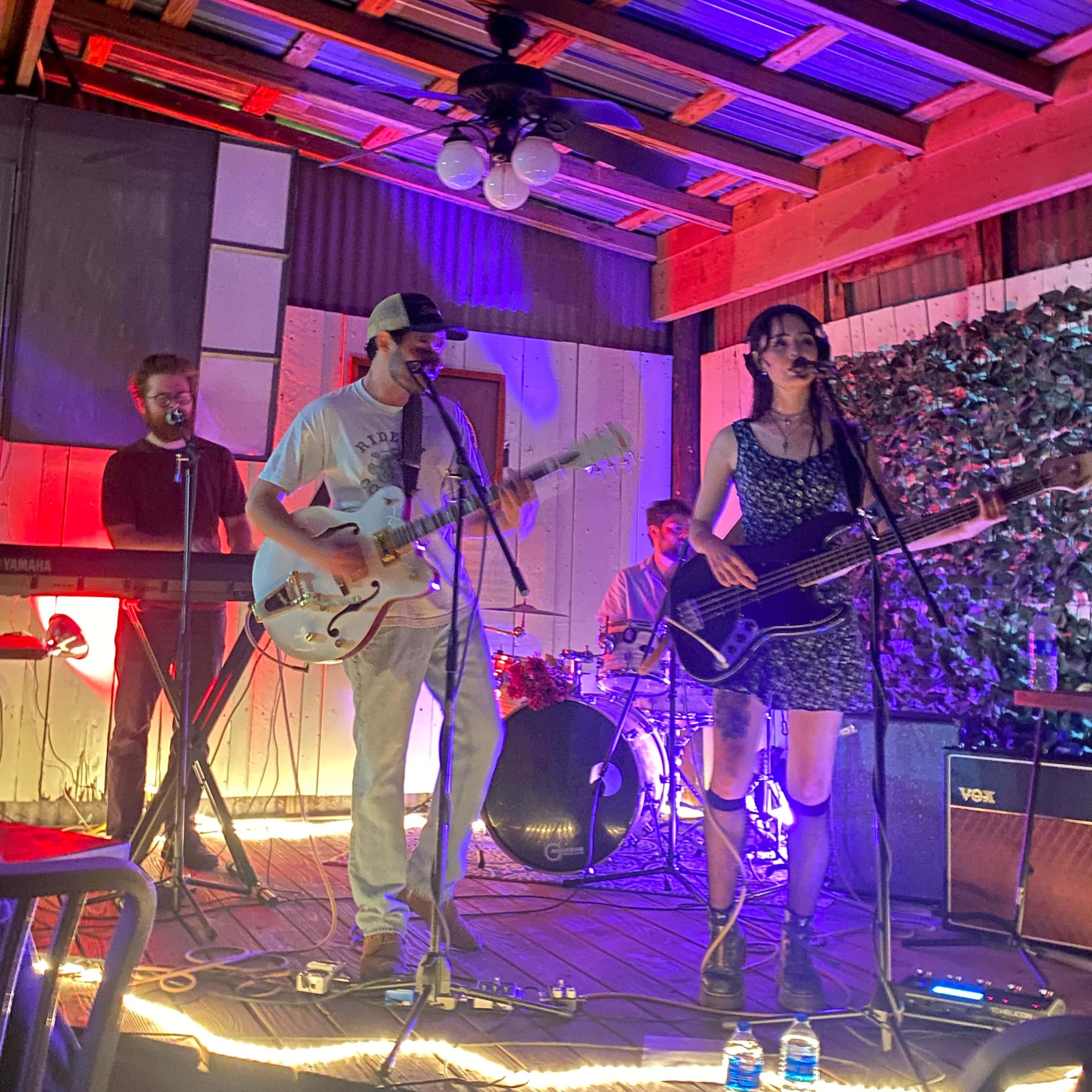 A band performs on a wooden stage under colorful lights, with a guitarist, bassist, drummer, and keyboardist present.