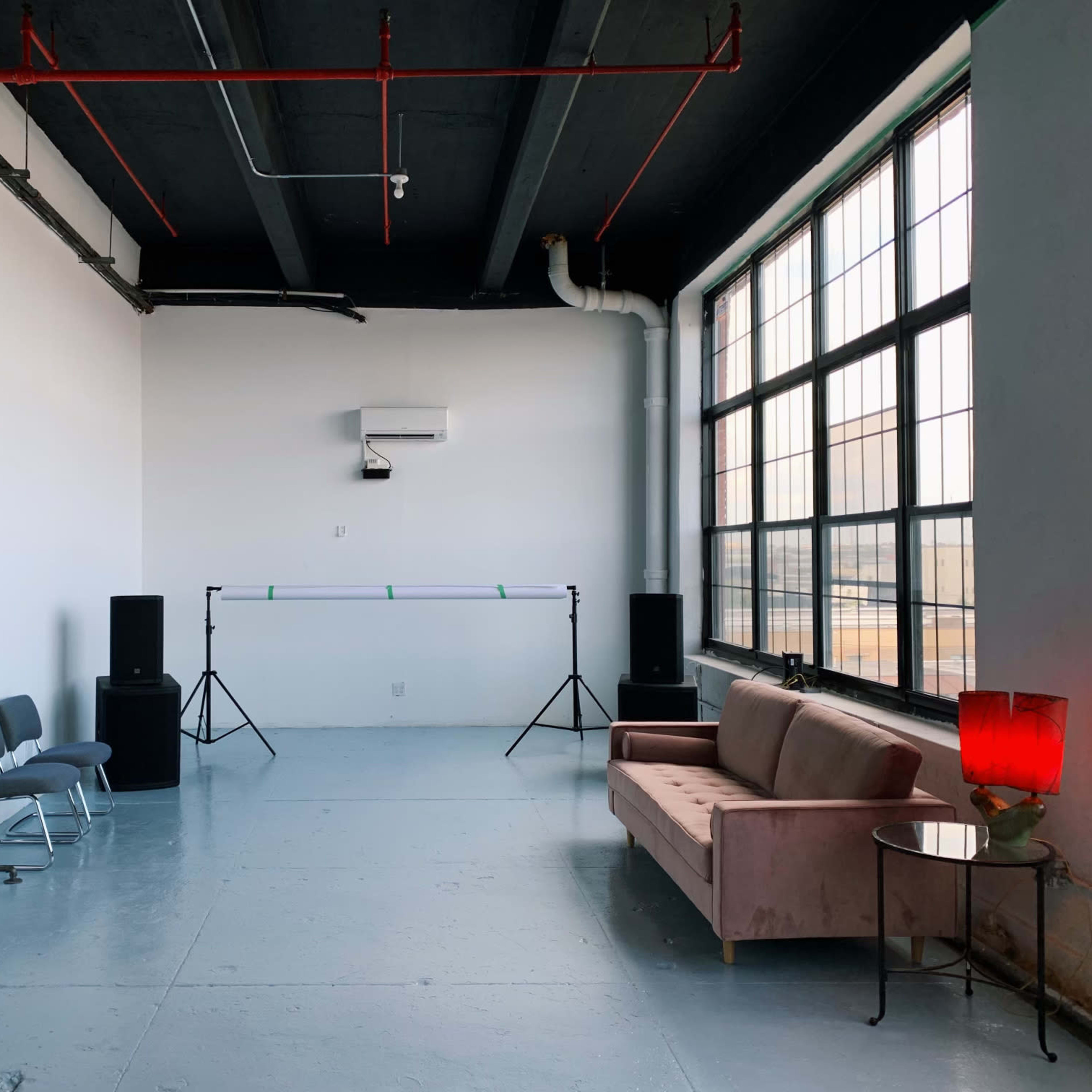 A minimalistic indoor space with a pink sofa, a side table with a red lamp, and audio equipment positioned beside a large window.