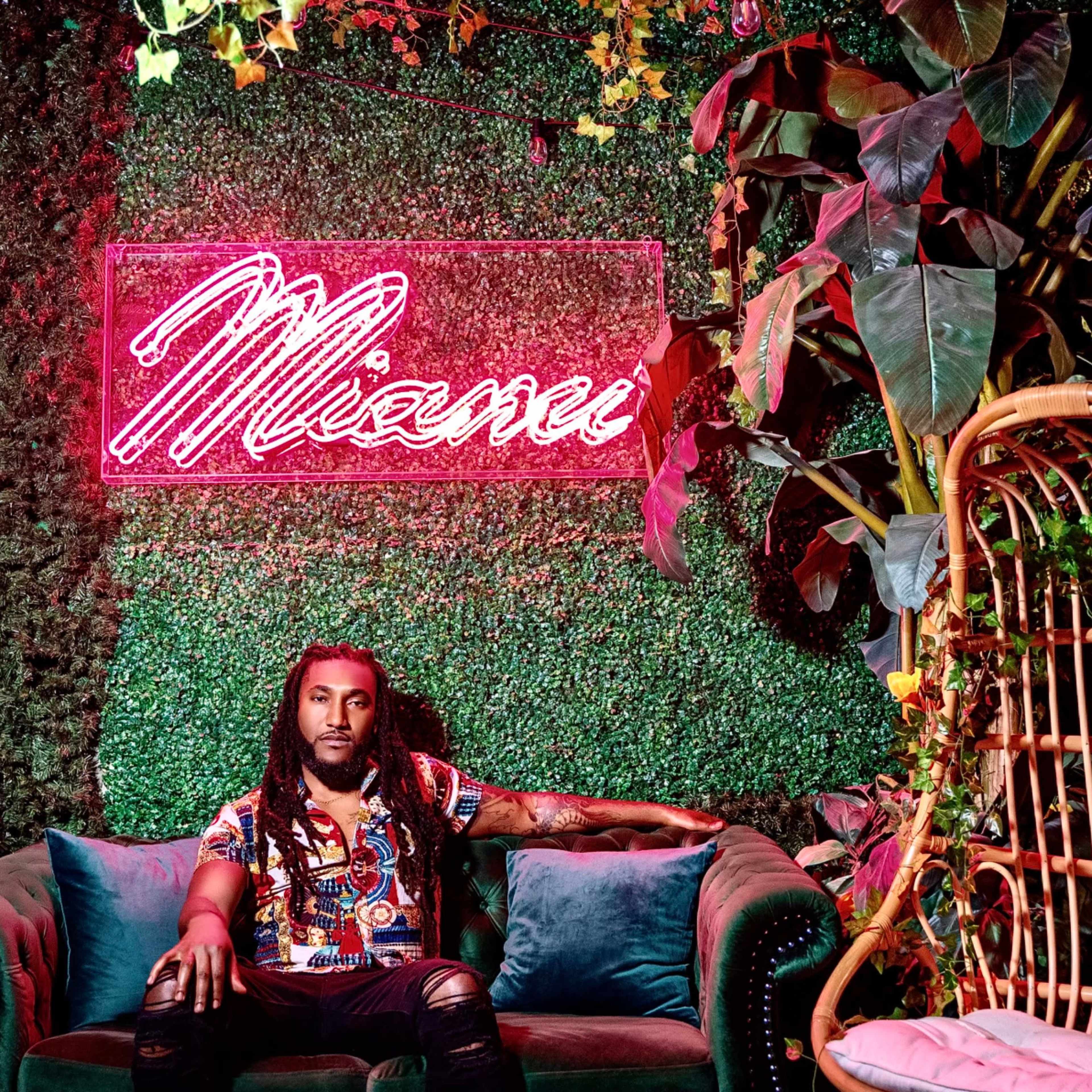 A man with long hair sits on a green sofa in front of a neon sign that says "Mienu," surrounded by lush plants and foliage.