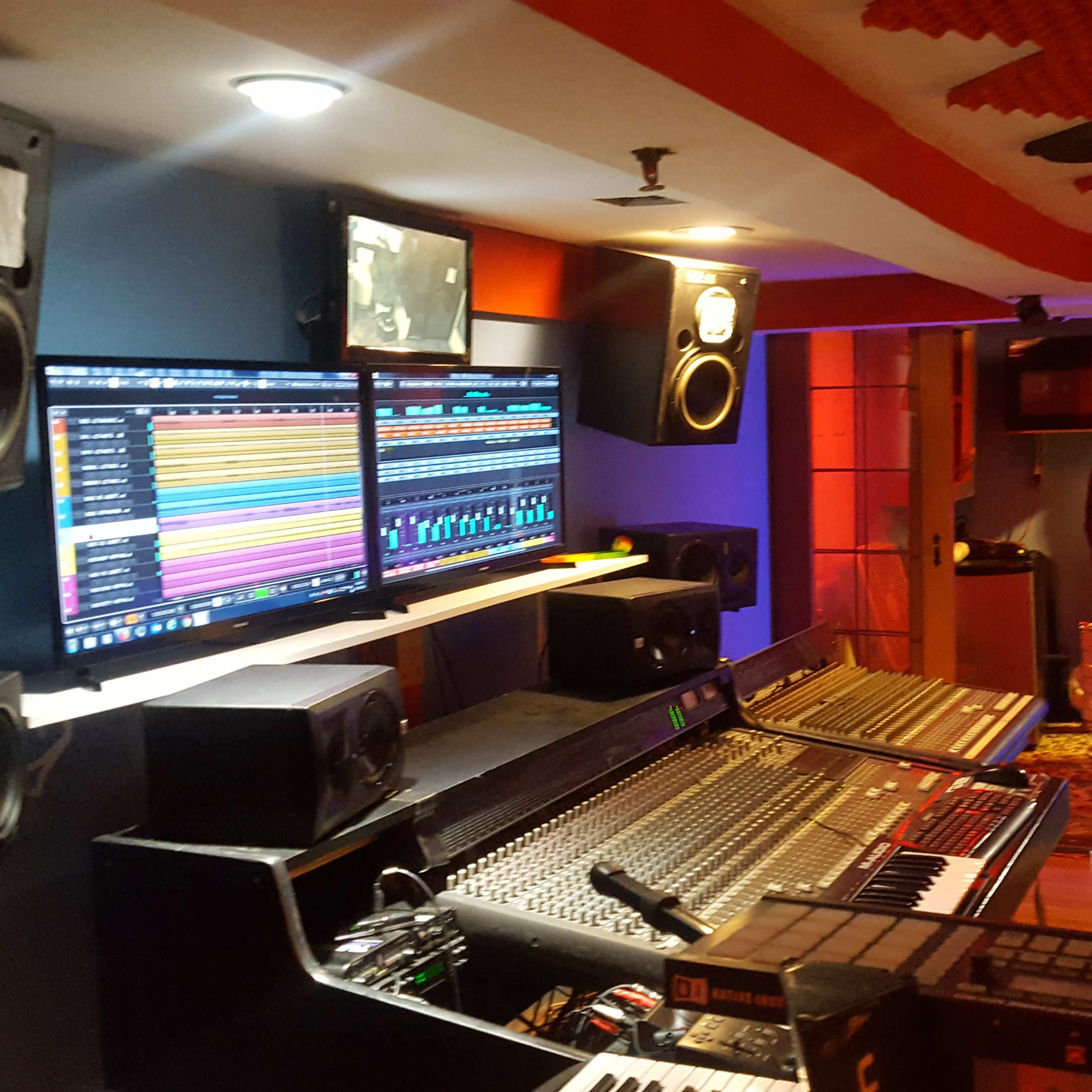 A modern recording studio with a mixing console, dual computer monitors displaying audio tracks, and speakers arranged around a slightly dimly lit room.