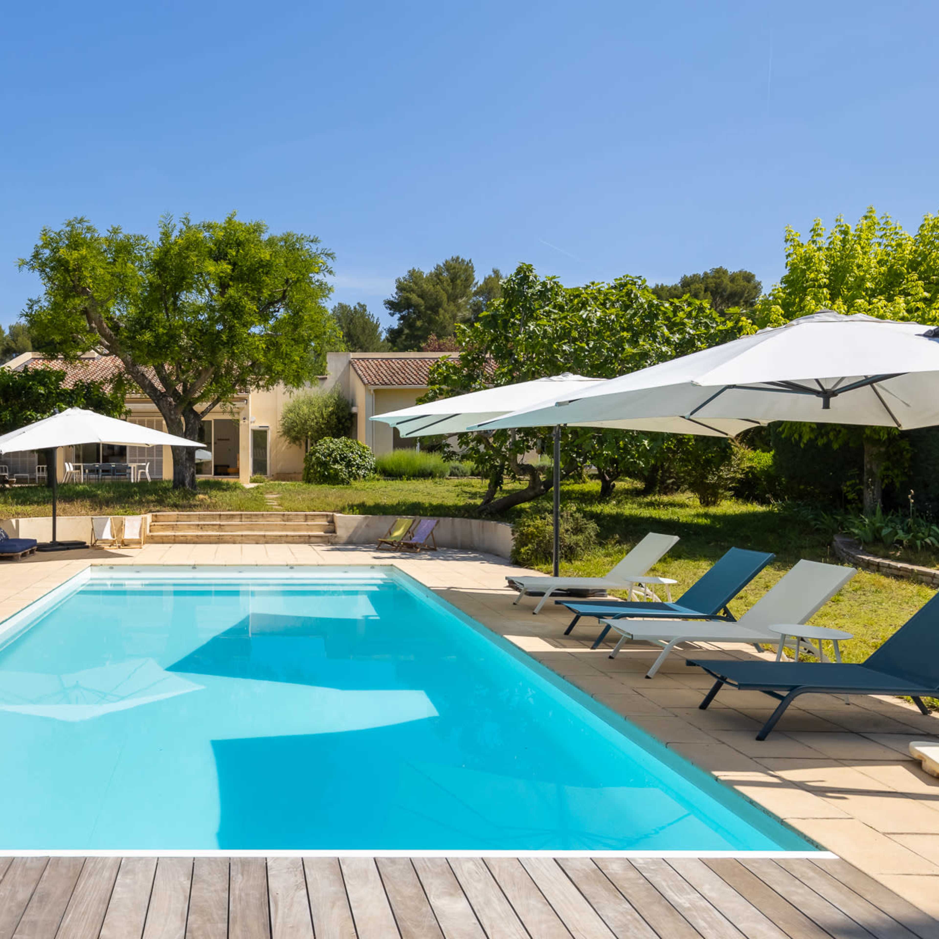 A clear swimming pool is surrounded by lounge chairs and parasols in a landscaped garden with trees and a house in the background.
