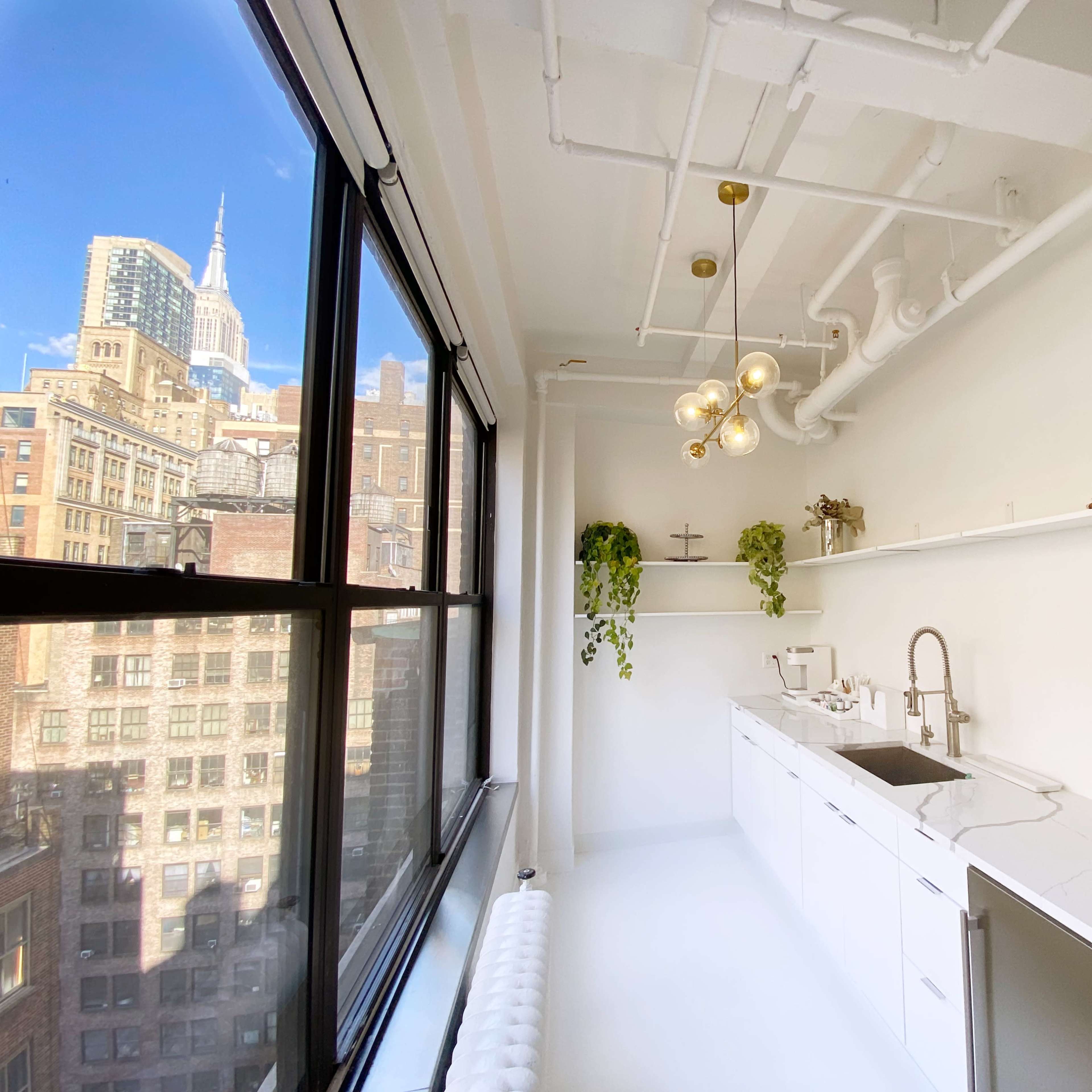 A modern kitchen with a white color scheme, large windows offering a view of the Empire State Building, and greenery adornment.
