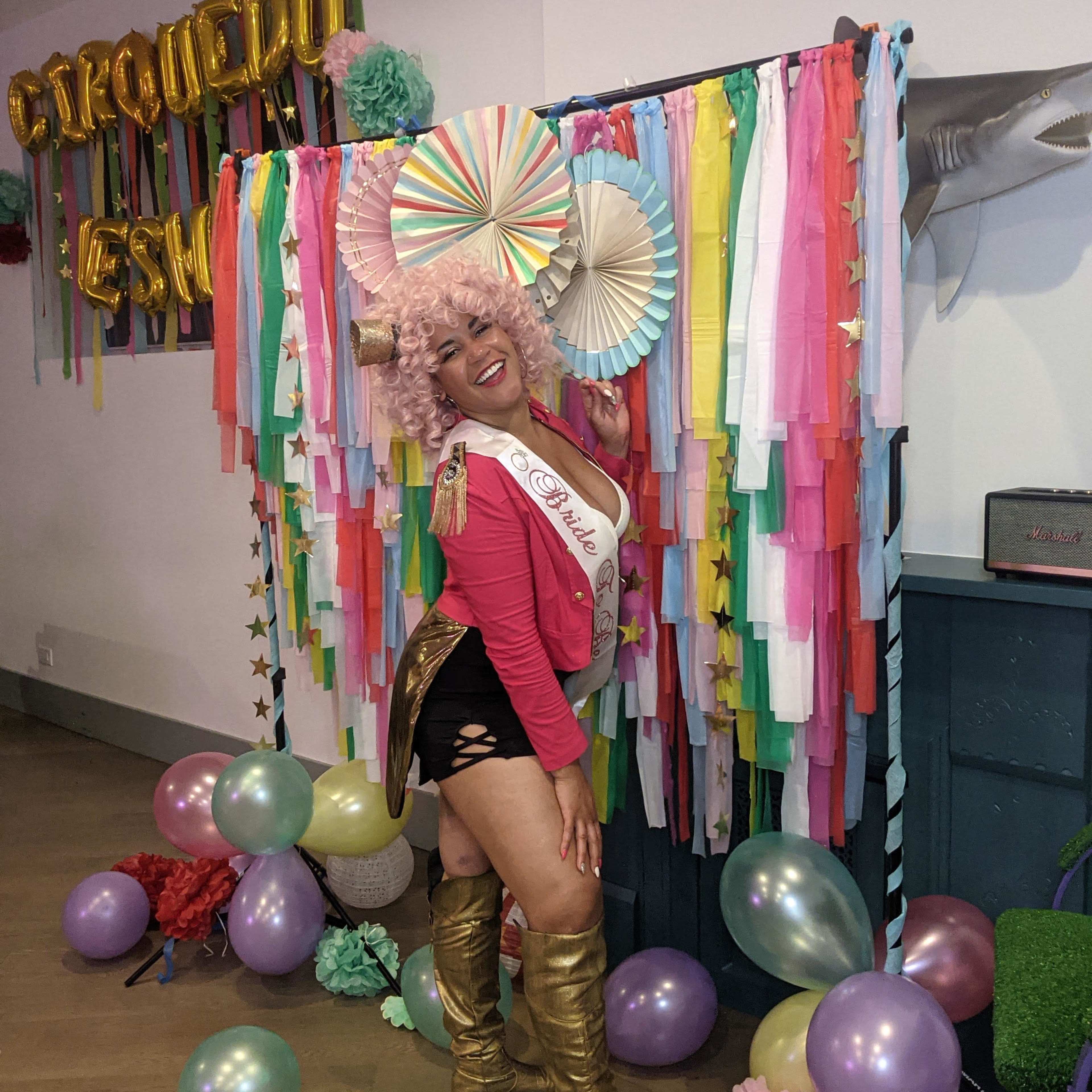 A person in a pink outfit and gold boots poses in front of a colorful backdrop decorated with streamers, paper fans, and balloons.
