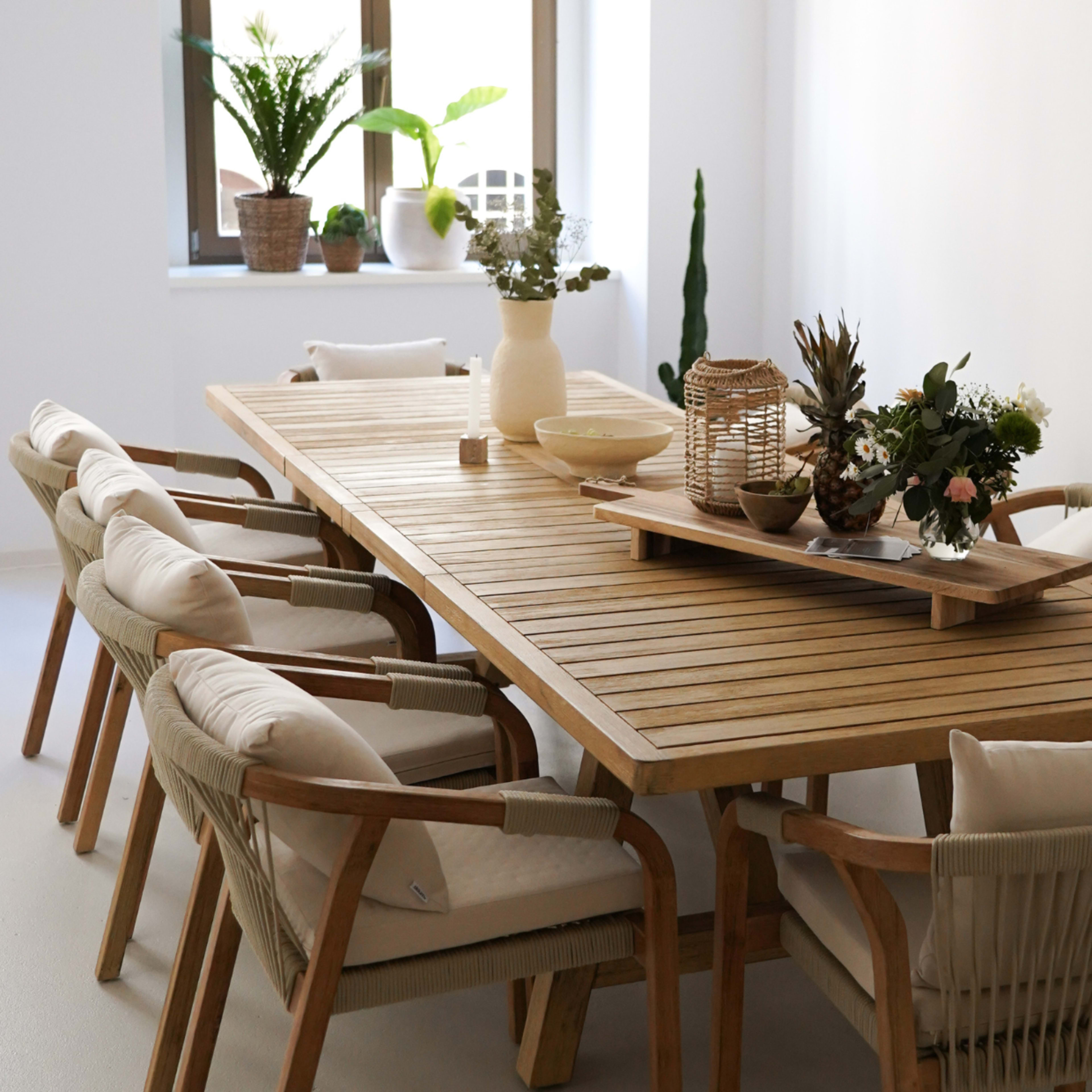 A wooden dining table with eight chairs, decorated with plants and a centerpiece, is set in a bright, minimalistic room.