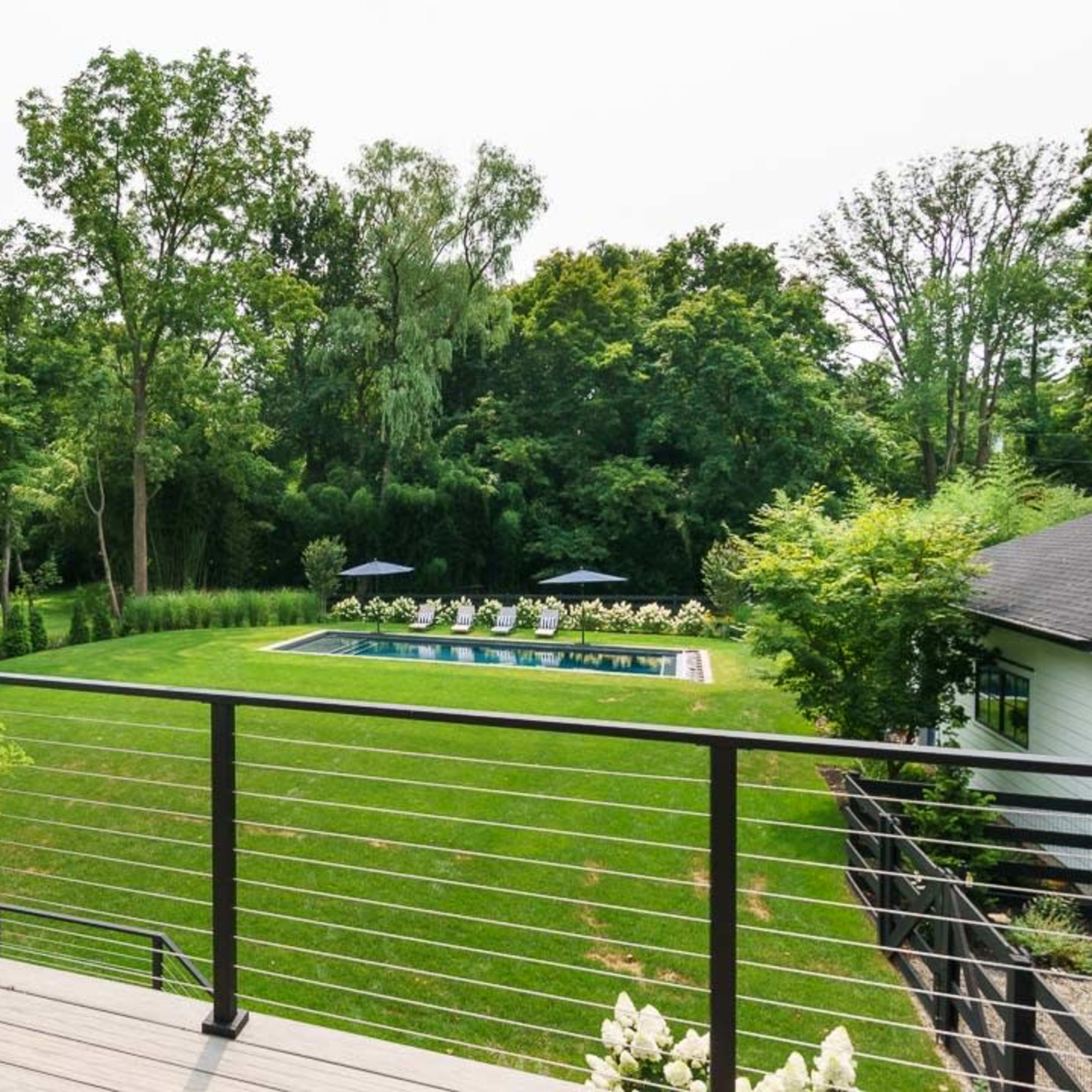 A deck overlooks a grassy area with a swimming pool surrounded by lounge chairs and trees.