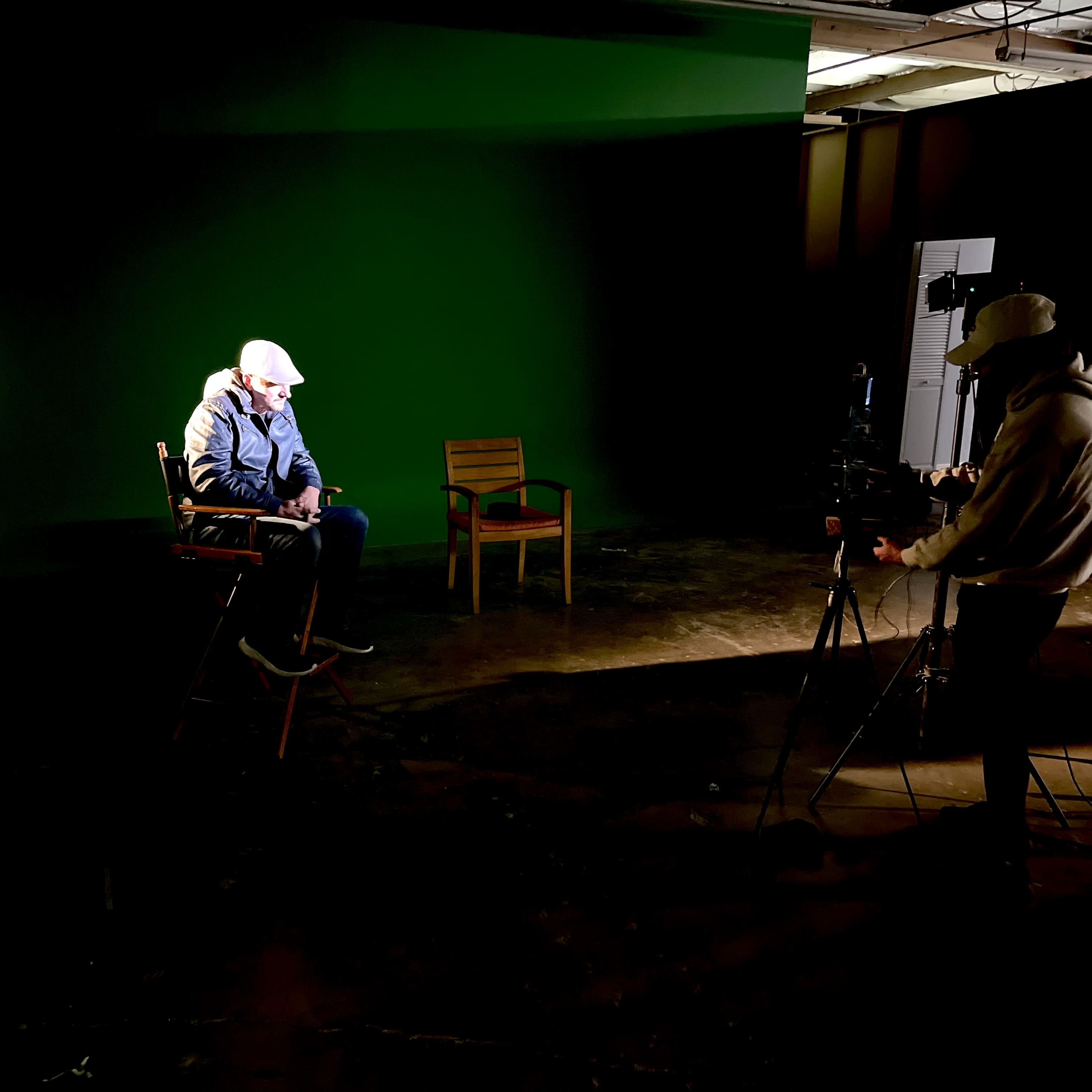 A person sits in a chair under spotlight on a green screen set while another person operates a camera.