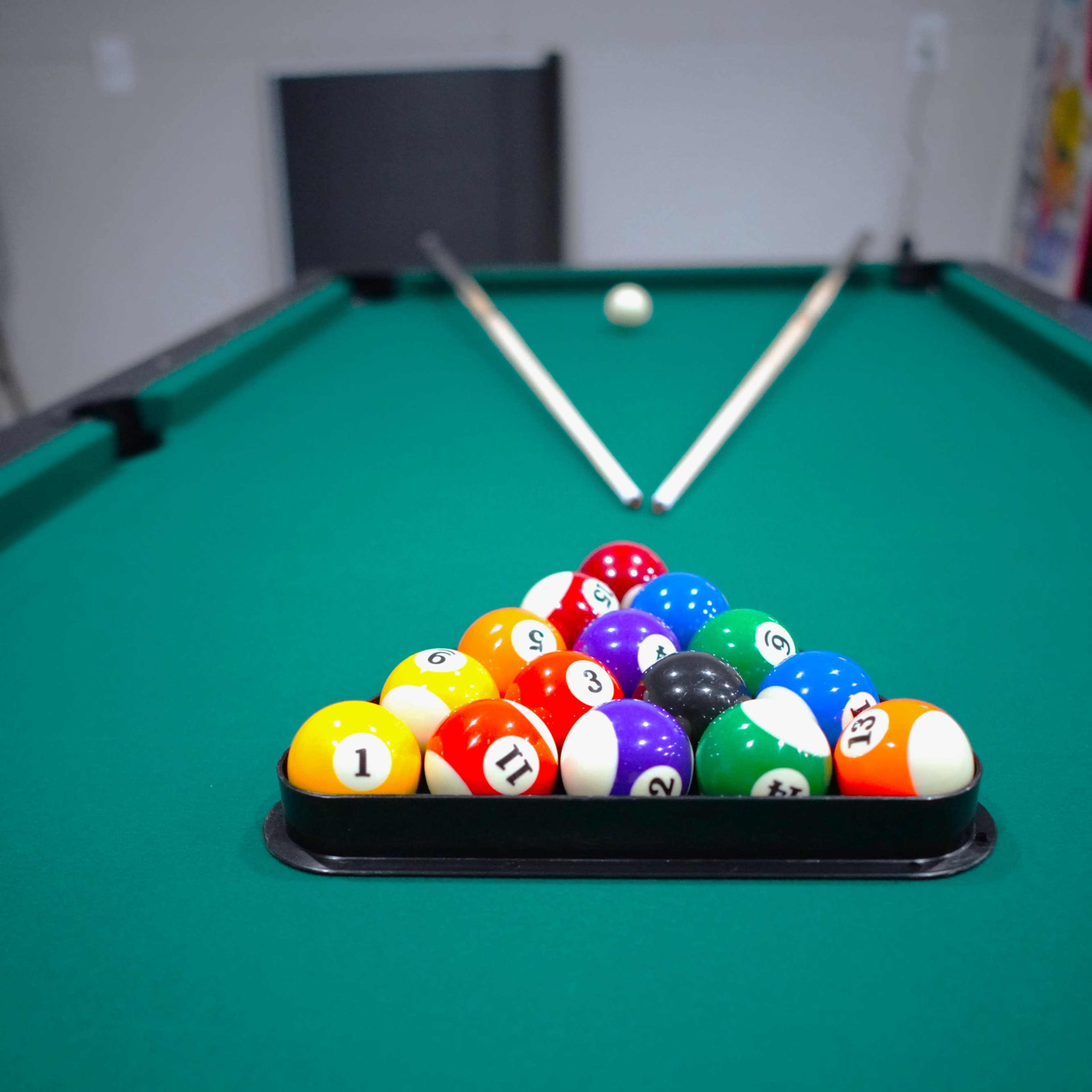 A triangle rack of colorful billiard balls is set on a green pool table, with two cue sticks resting beside it.