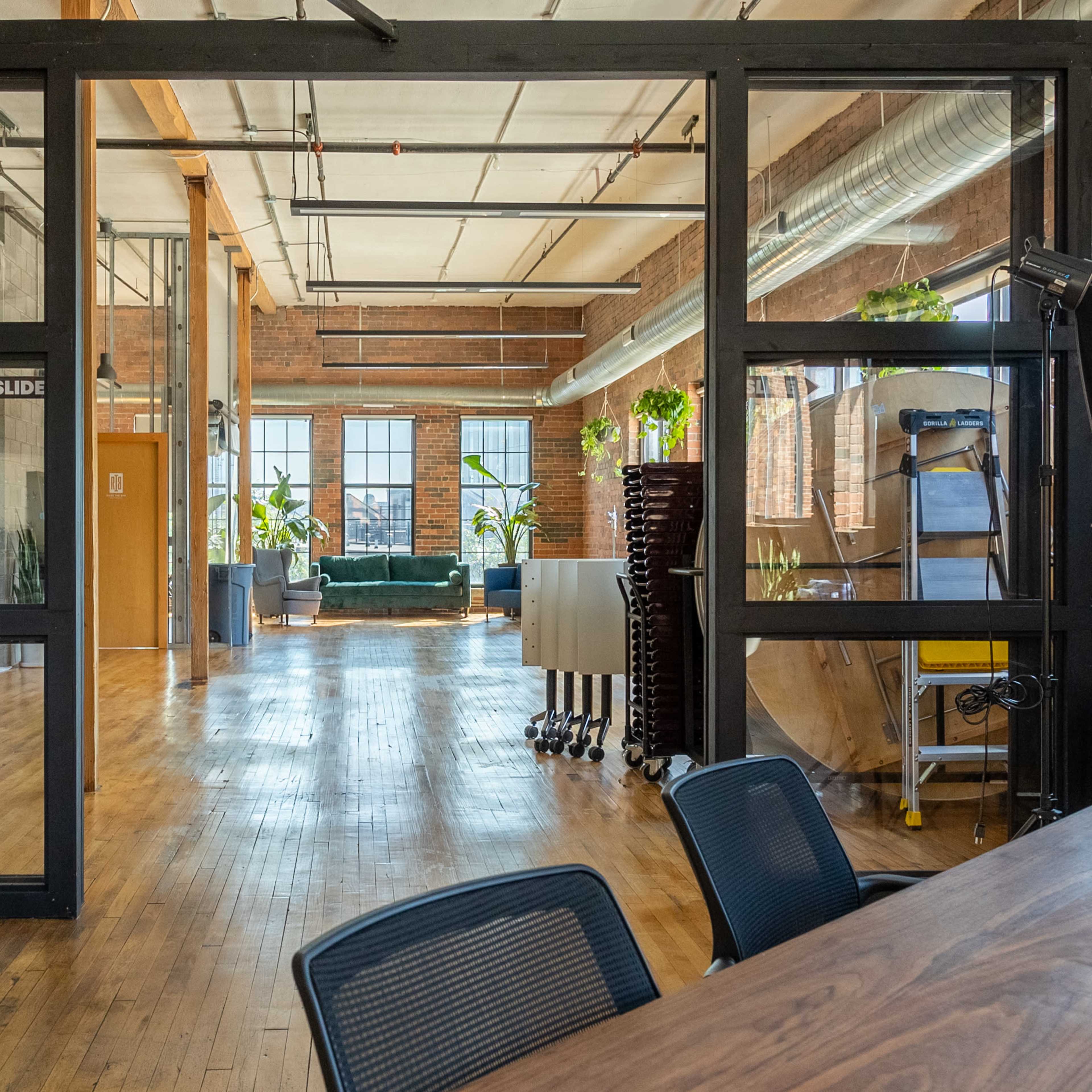 A spacious, modern office interior with large windows, wooden floors, and various workspaces separated by glass walls.
