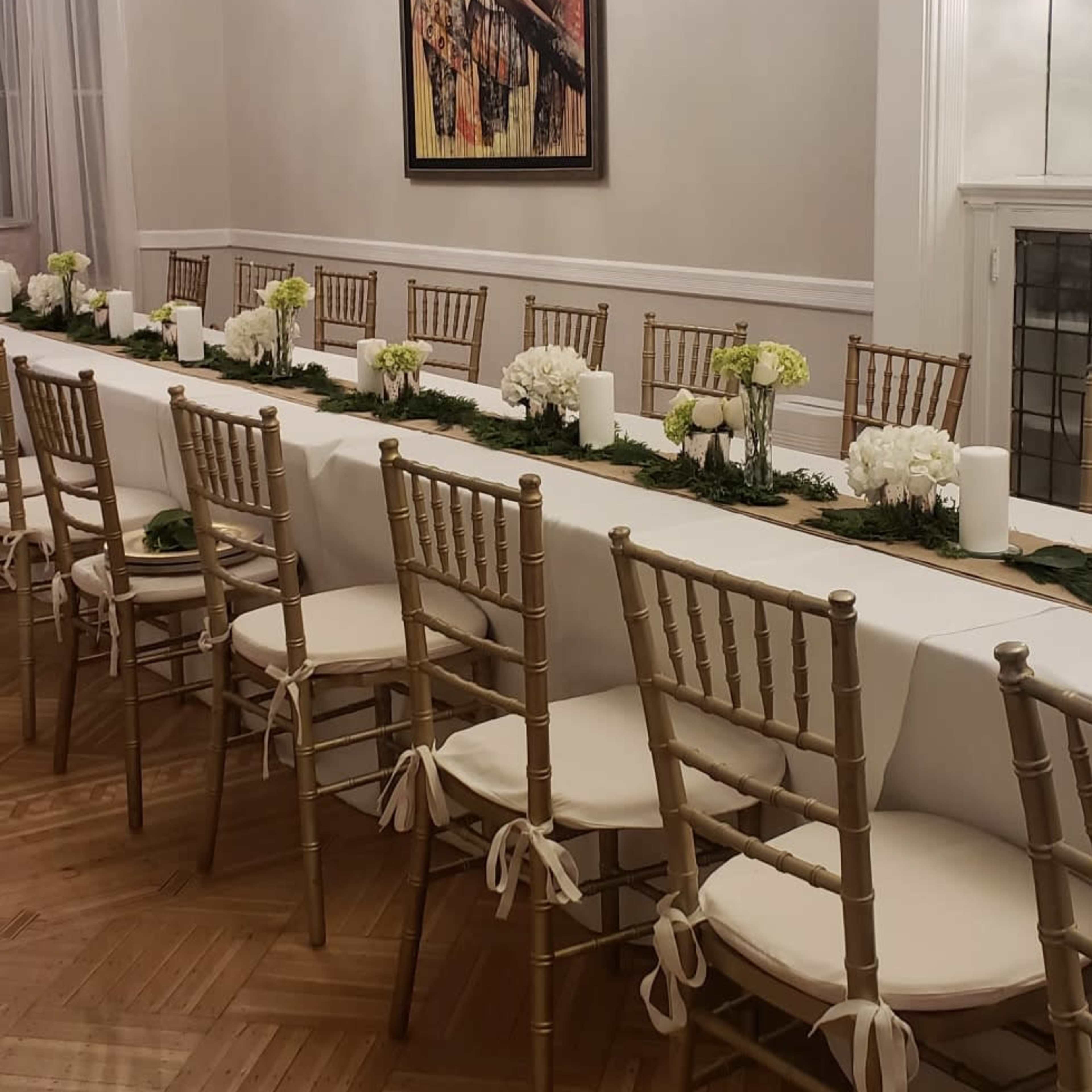 A long dining table is set with white linens and floral centerpieces in a well-lit room featuring a decorative chandelier and an abstract painting on the wall.
