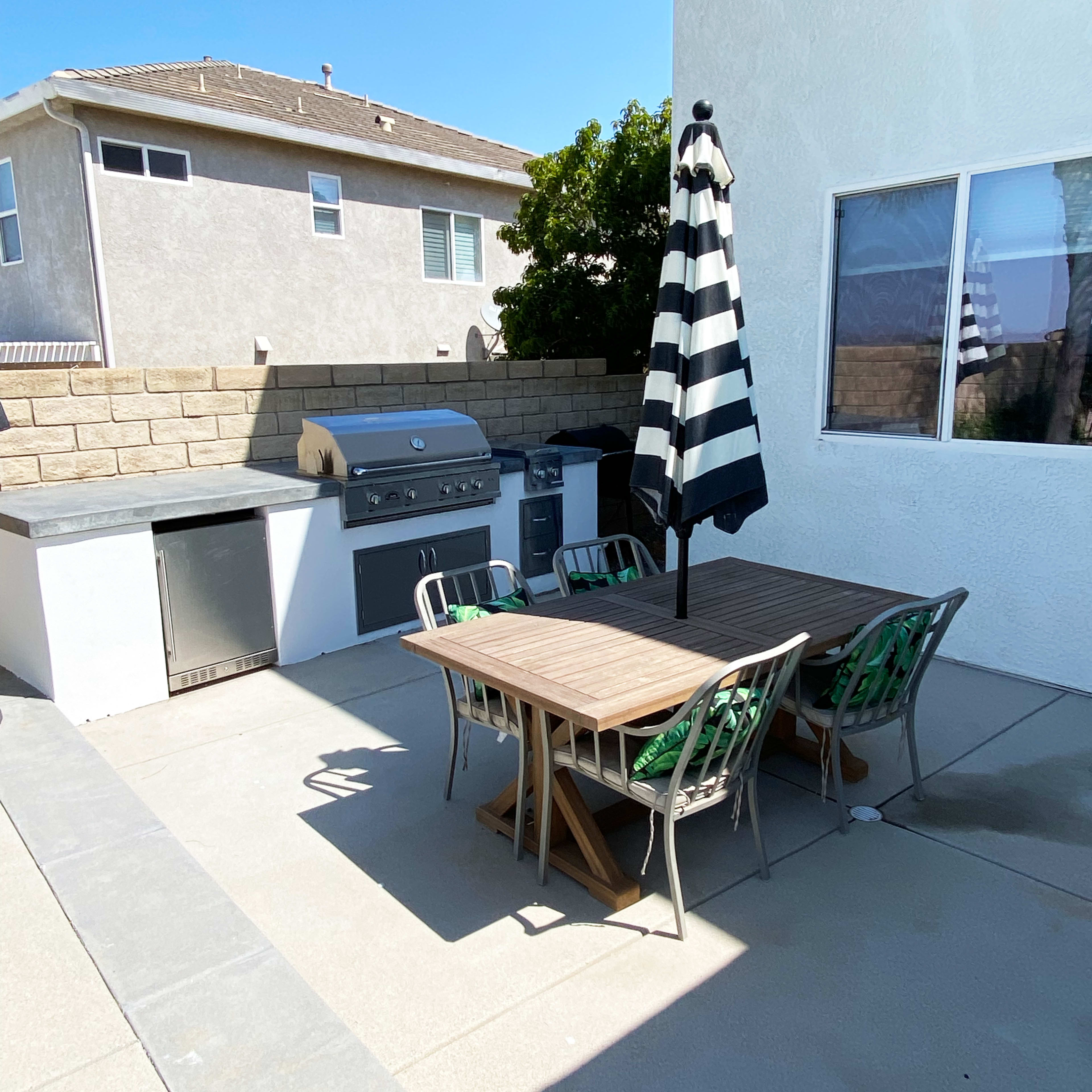 An outdoor patio area with a table and chairs, a grill, and an umbrella, surrounded by a concrete surface and a low wall.