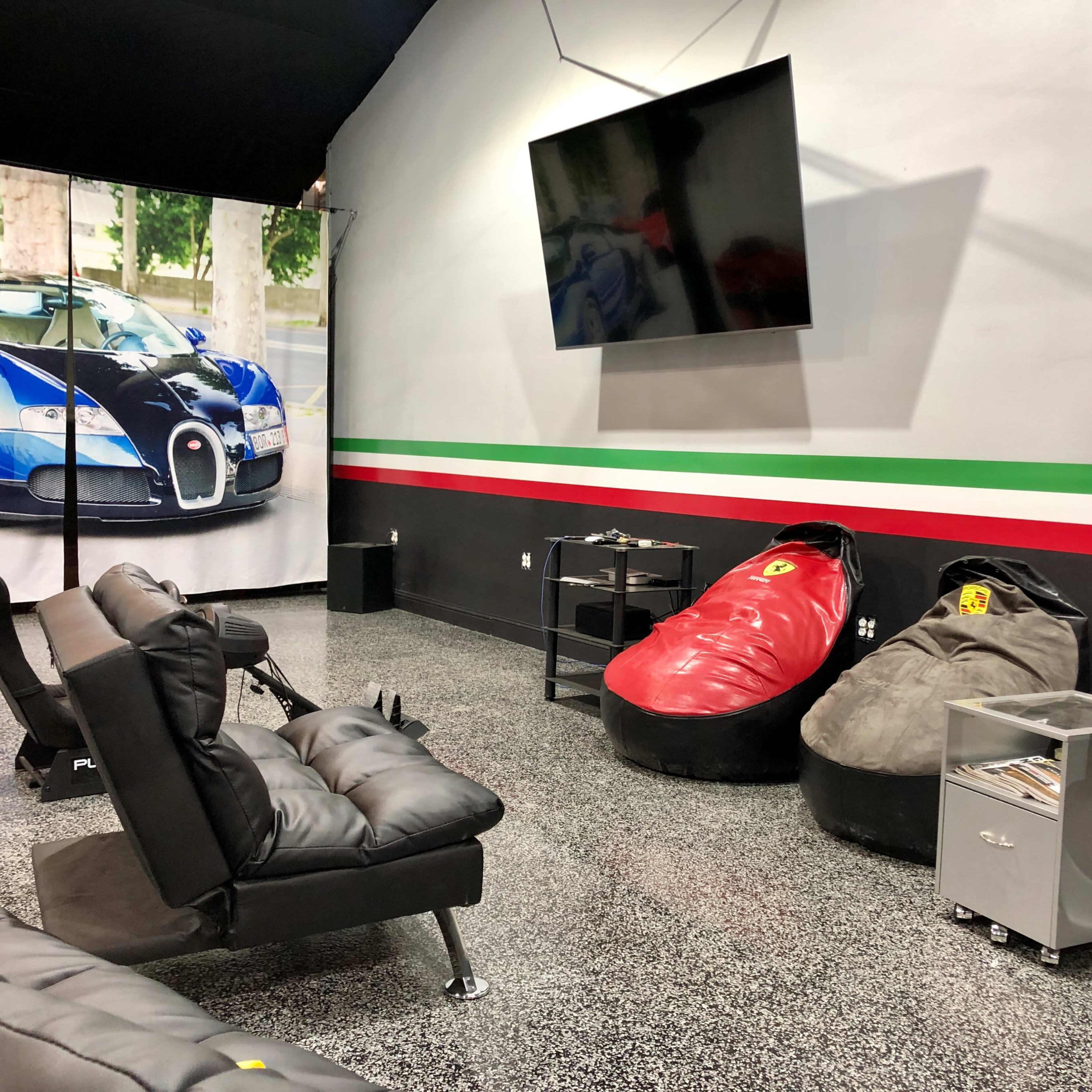 A modern lounge area featuring black leather seating, bean bags resembling sports cars, and a large flat-screen TV mounted on the wall, with a backdrop of a luxury car poster.