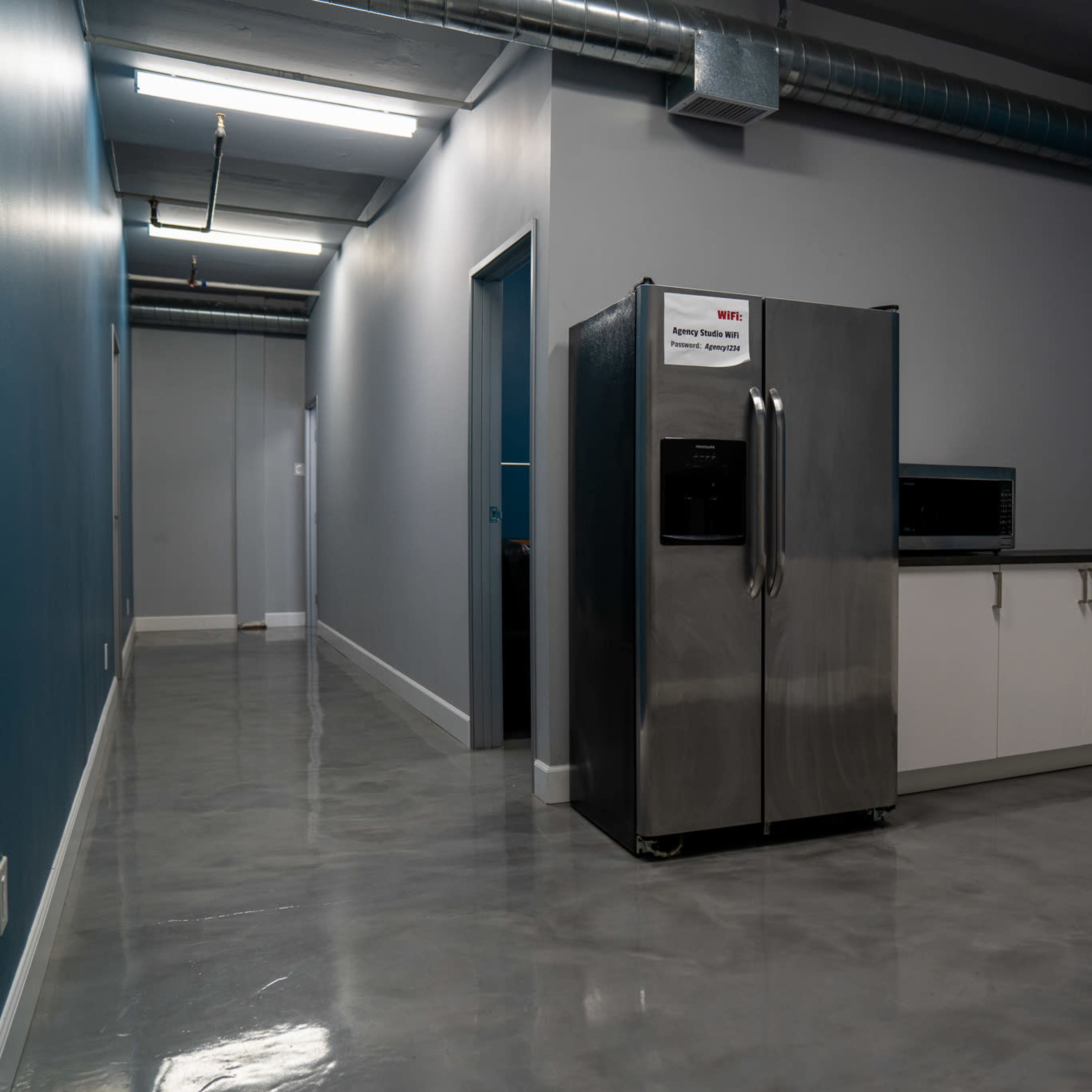 A kitchen area with a stainless steel refrigerator and a long countertop, adjacent to a hallway with gray walls and a blue accent wall.