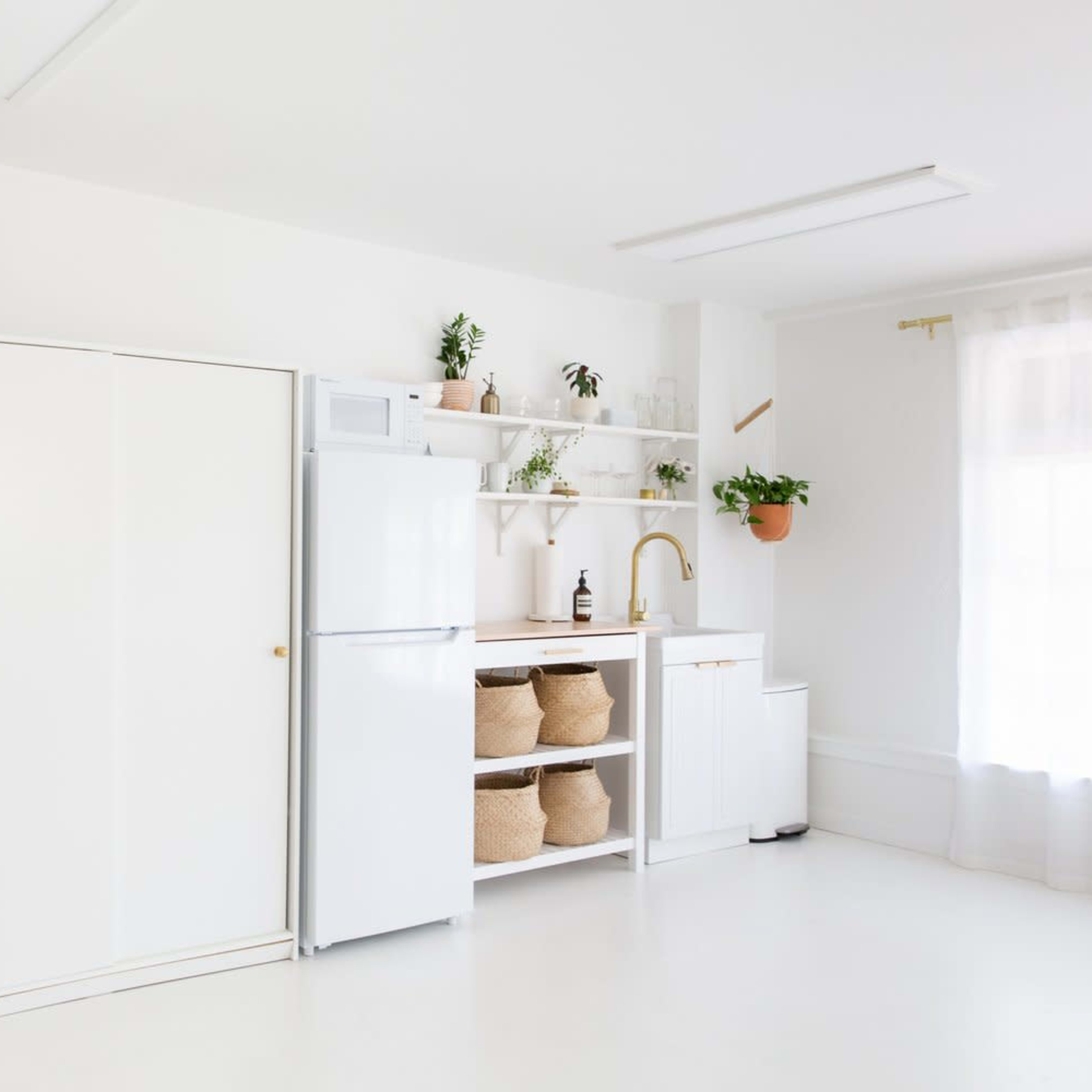 A modern kitchen features white cabinetry, a refrigerator, open shelving with plants, and natural light streaming through large windows.