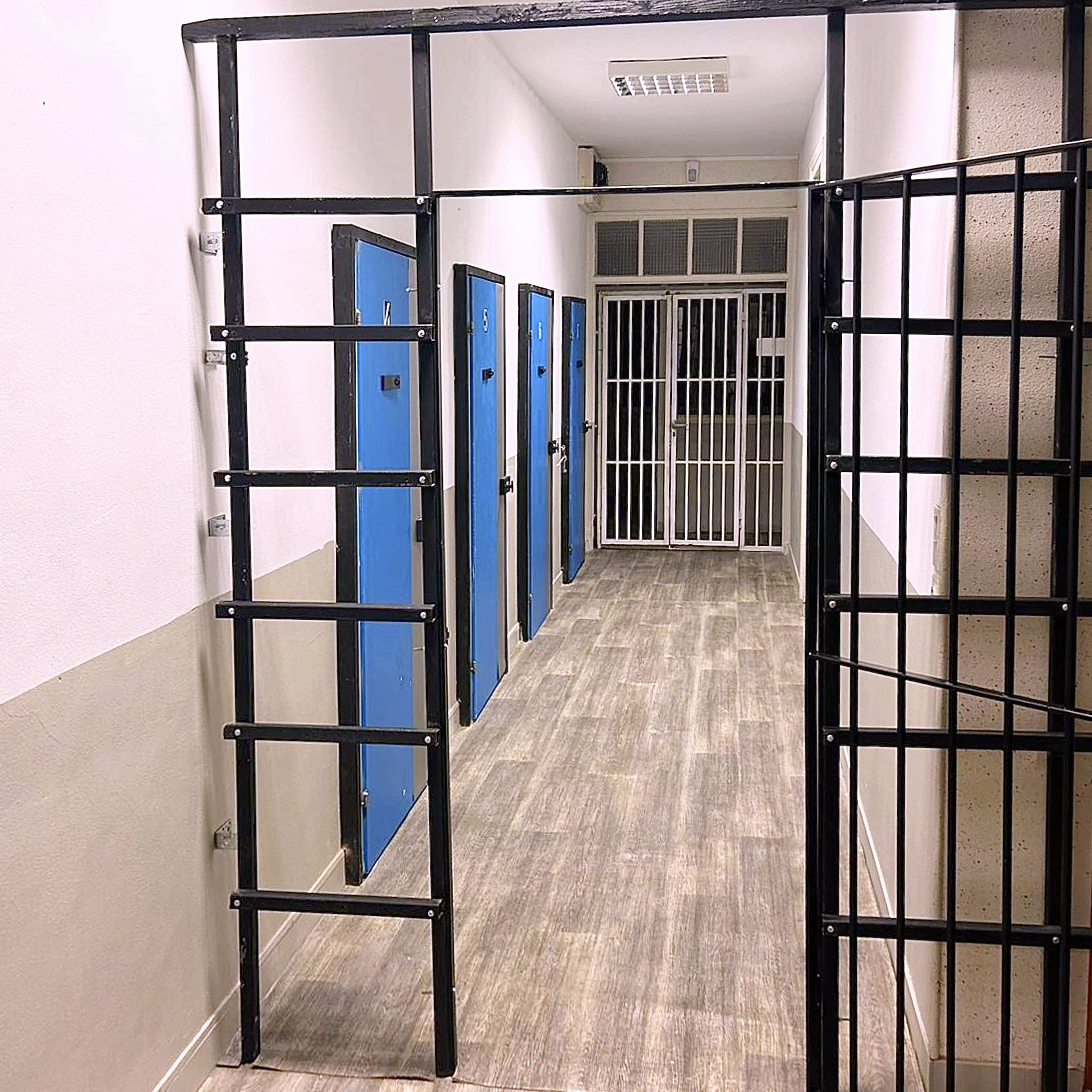 A narrow hallway in a prison with blue cell doors and a barred gate at the entrance.