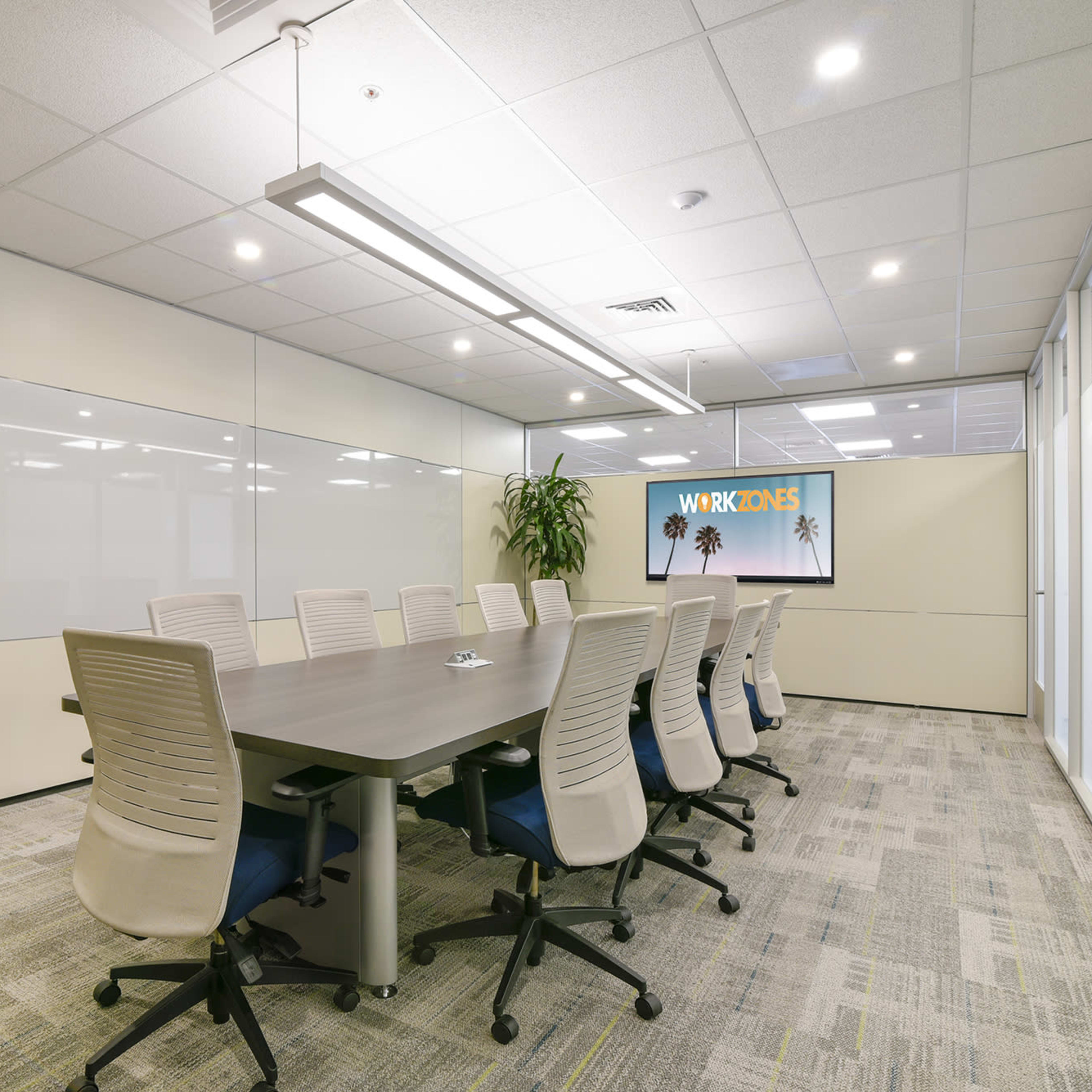 A modern conference room features a long table surrounded by ergonomic chairs, with a large screen displaying "WORKZONES" and glass walls allowing natural light.