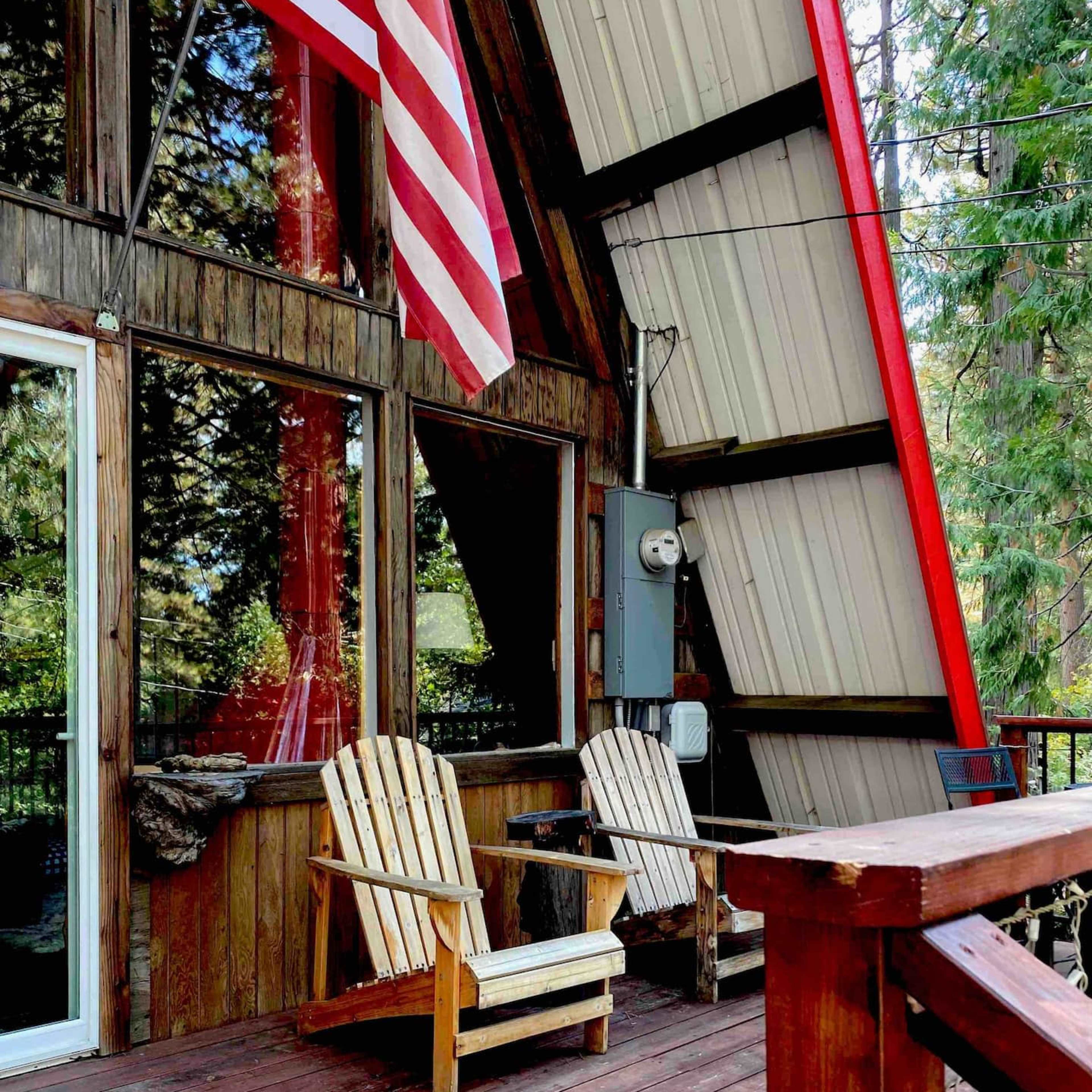 A wooden deck with two Adirondack chairs and an American flag displayed on a cabin-like structure surrounded by trees.