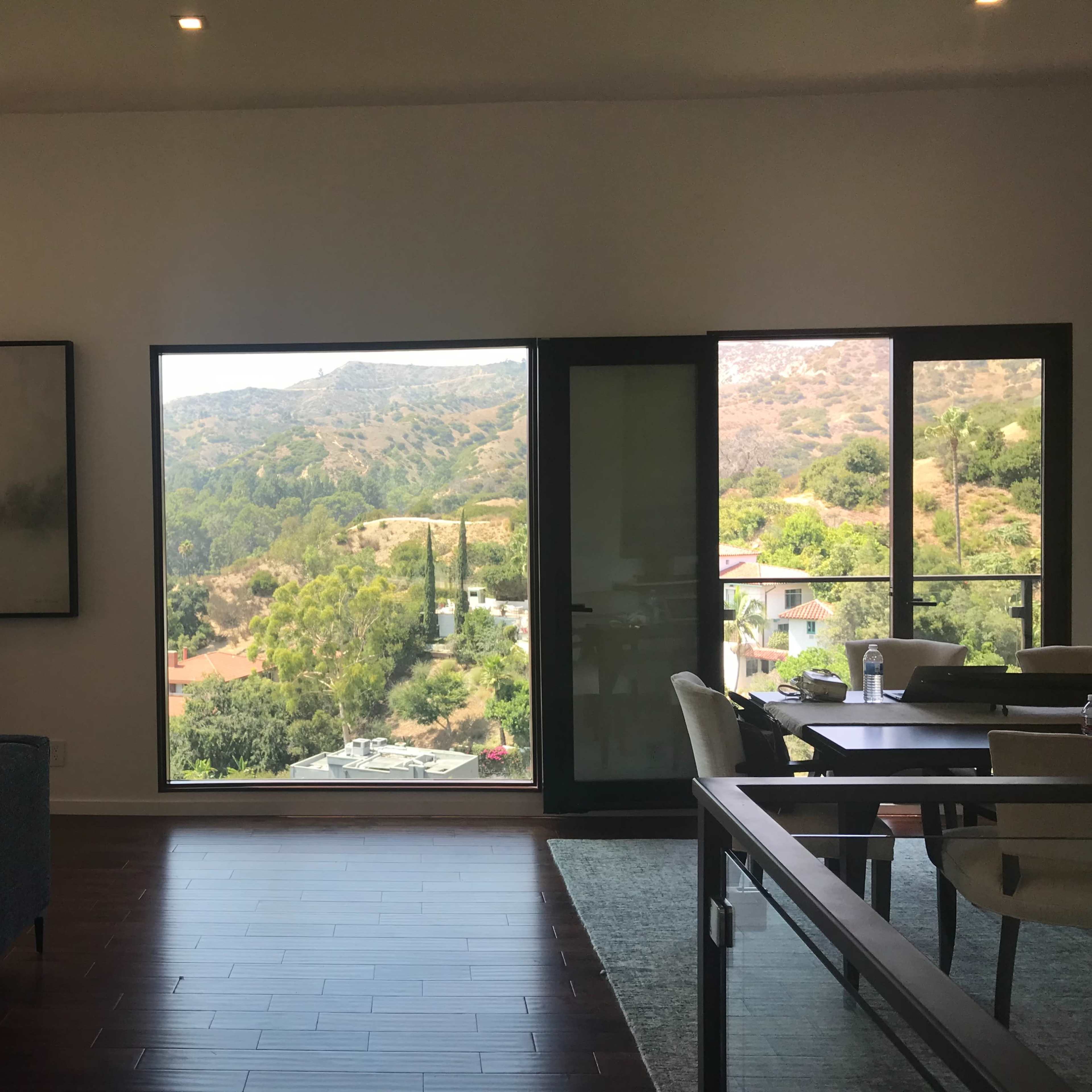 A modern living room with large windows overlooking a green hillside and adjacent buildings.