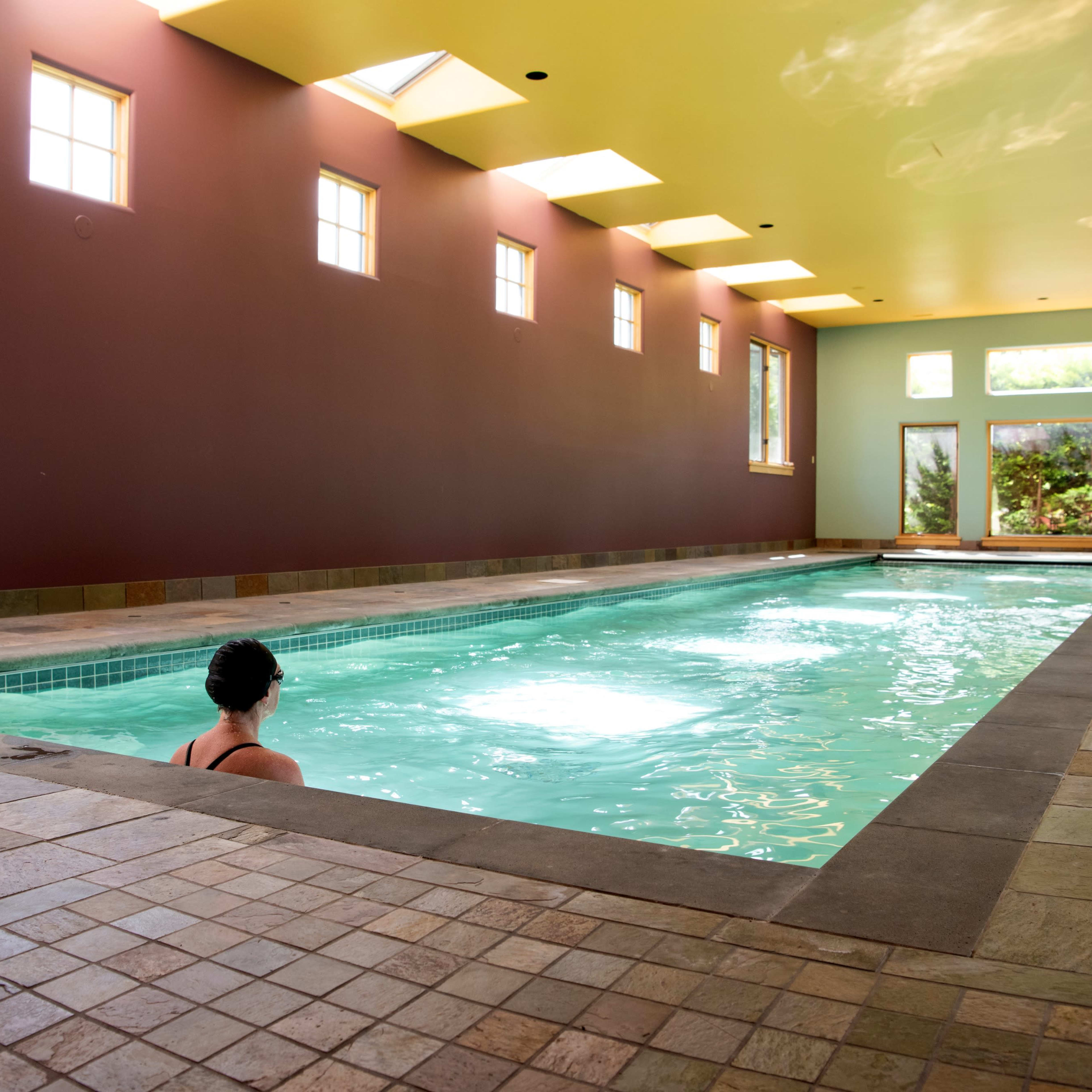 A person sits at the edge of an indoor swimming pool surrounded by large windows and brightly colored walls.
