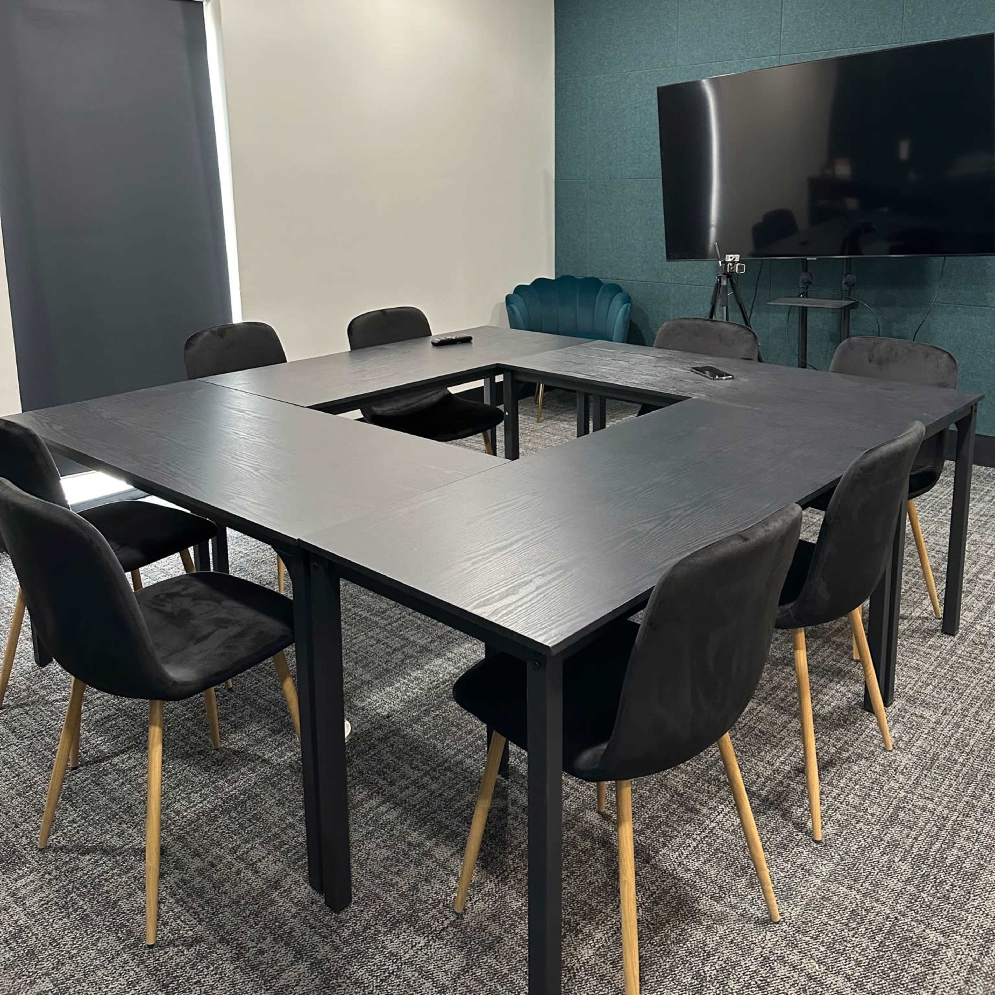 A modern conference room features a rectangular table surrounded by black chairs, with a large screen on one wall and a green accent wall.