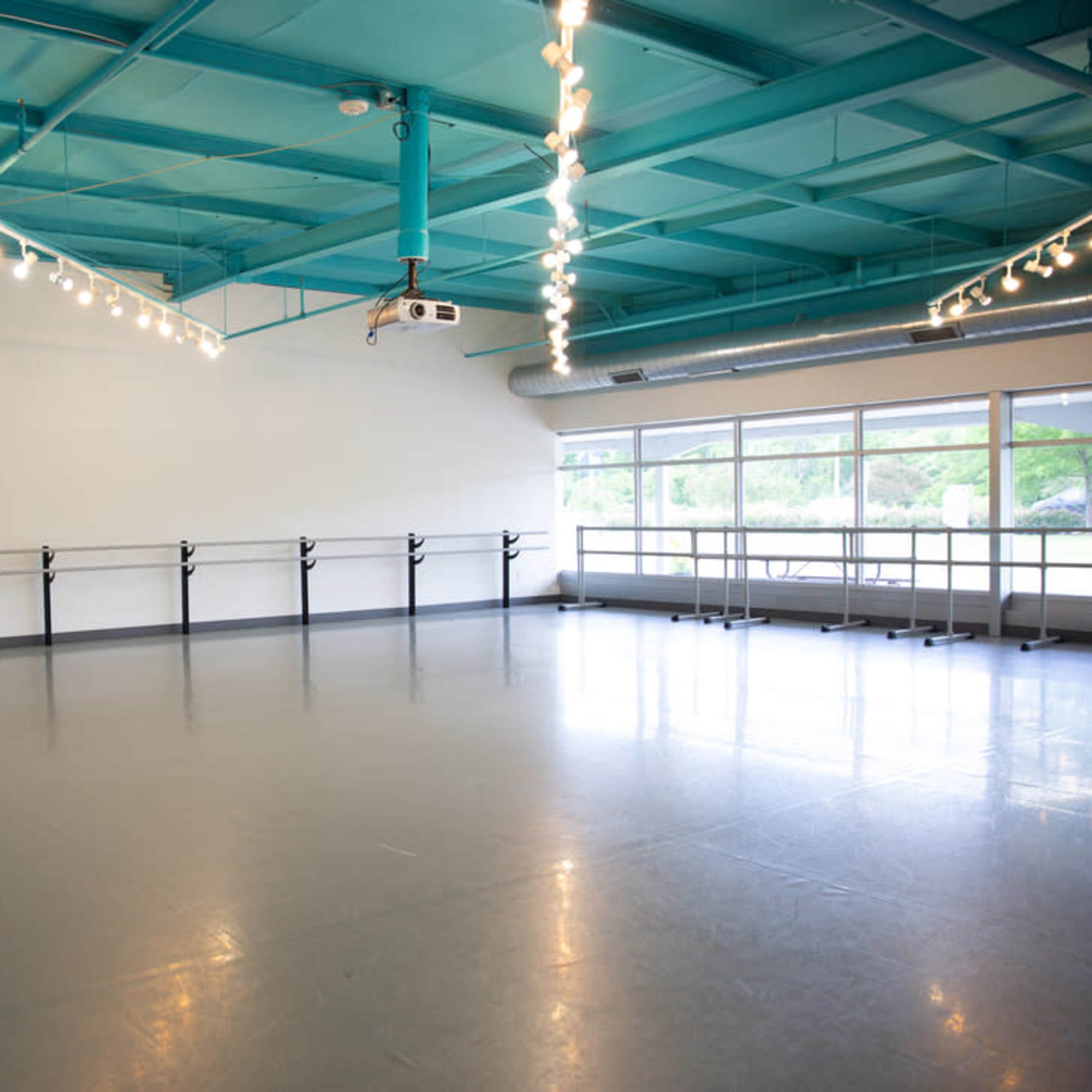 A spacious dance studio with a smooth gray floor, a turquoise ceiling, and large windows allowing natural light.