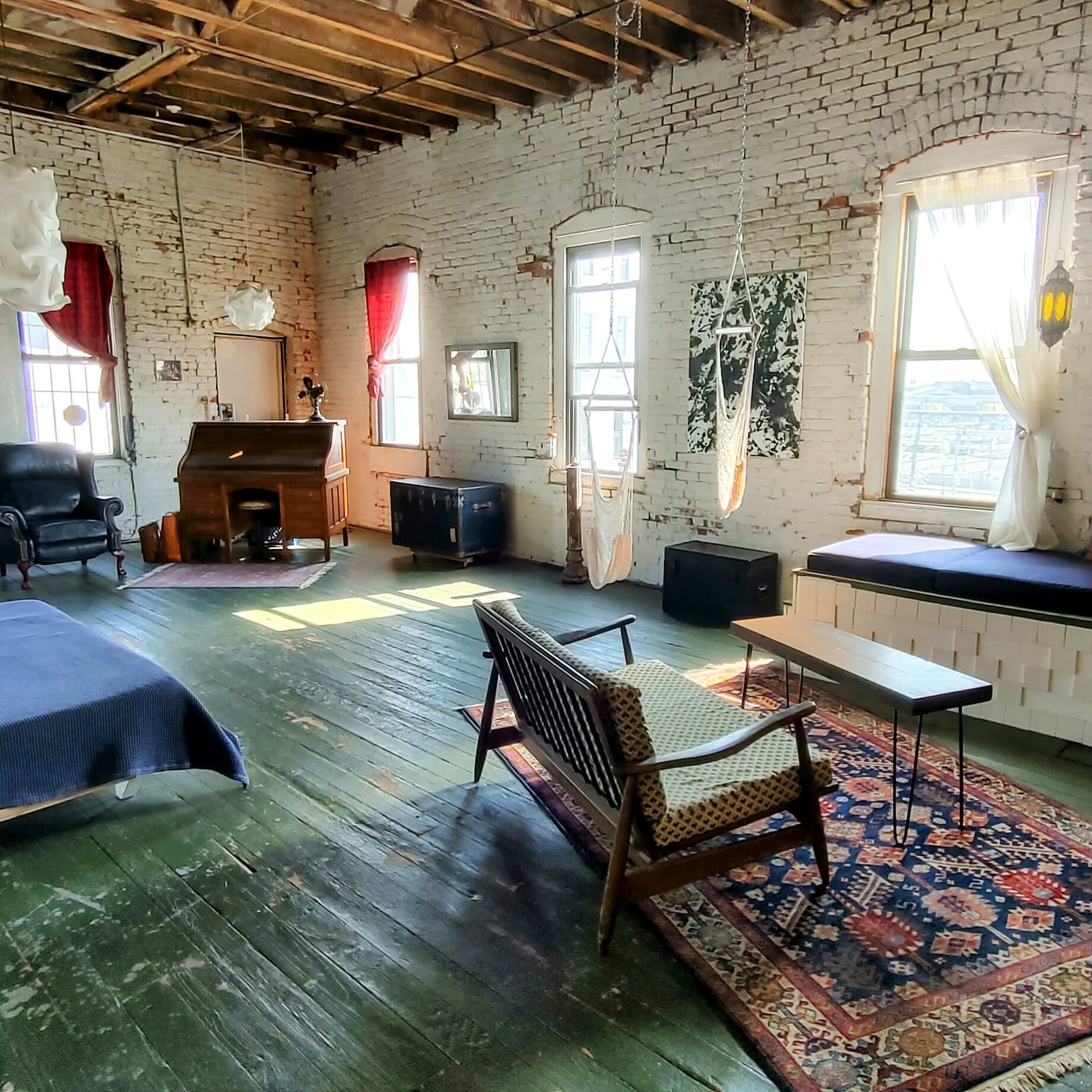 A spacious, rustic bedroom with exposed brick walls, large windows, and a mix of vintage furniture including a bed, a black chair, a wooden piano, and a patterned rug.