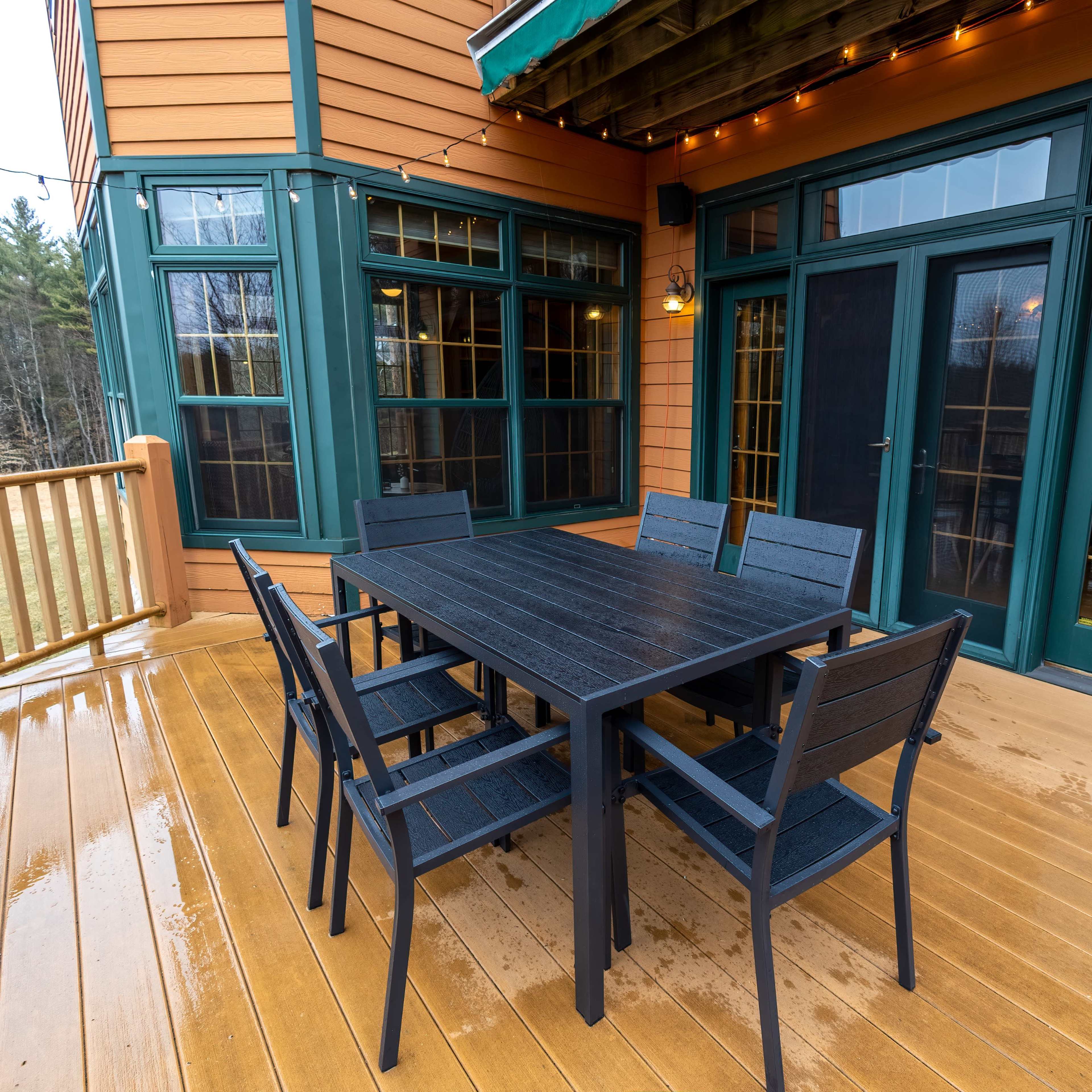 A dark wooden dining table with six black chairs is set on a wooden deck outside a house with green trim and large glass doors.
