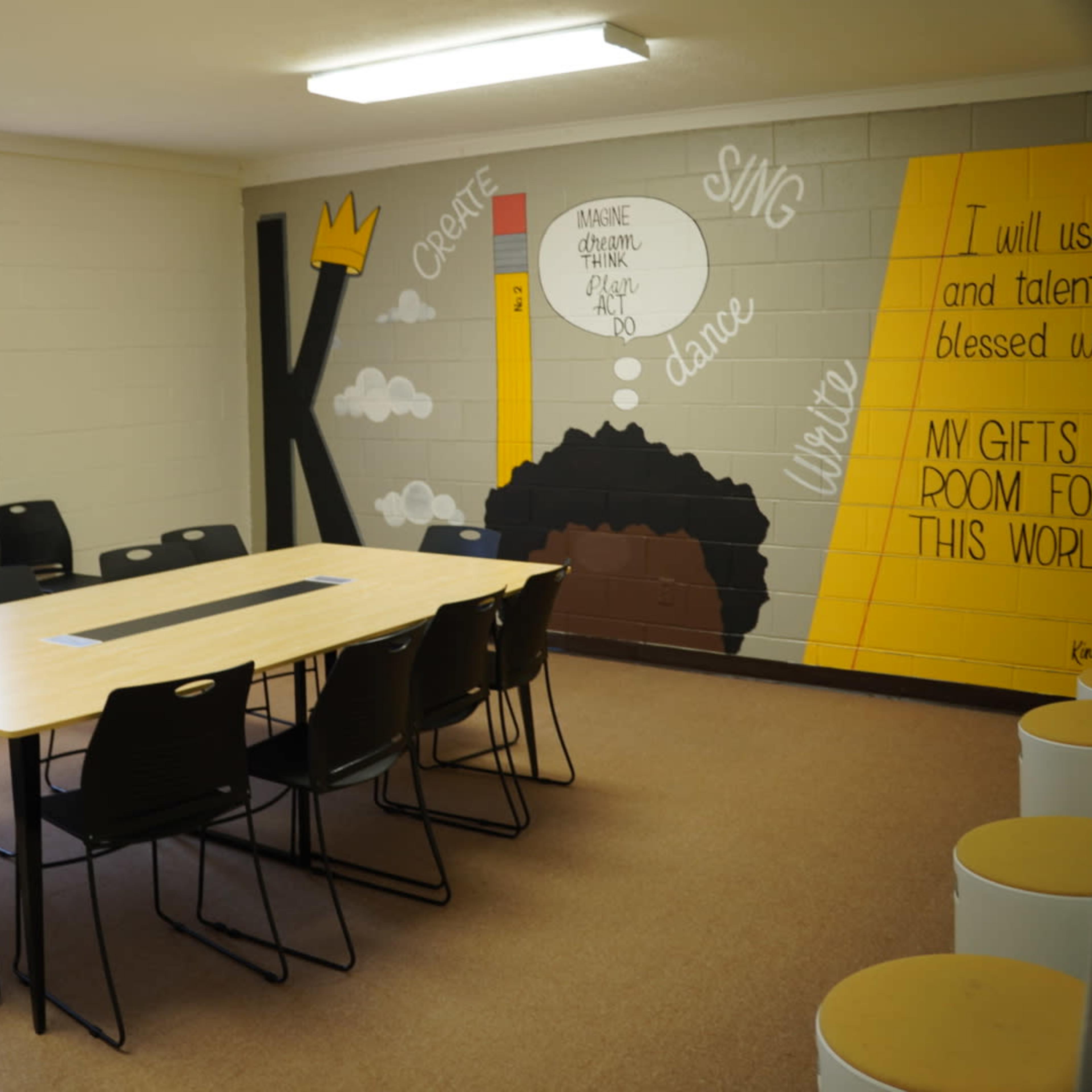 A classroom with a large table surrounded by black chairs and a colorful mural on the wall featuring motivational phrases.