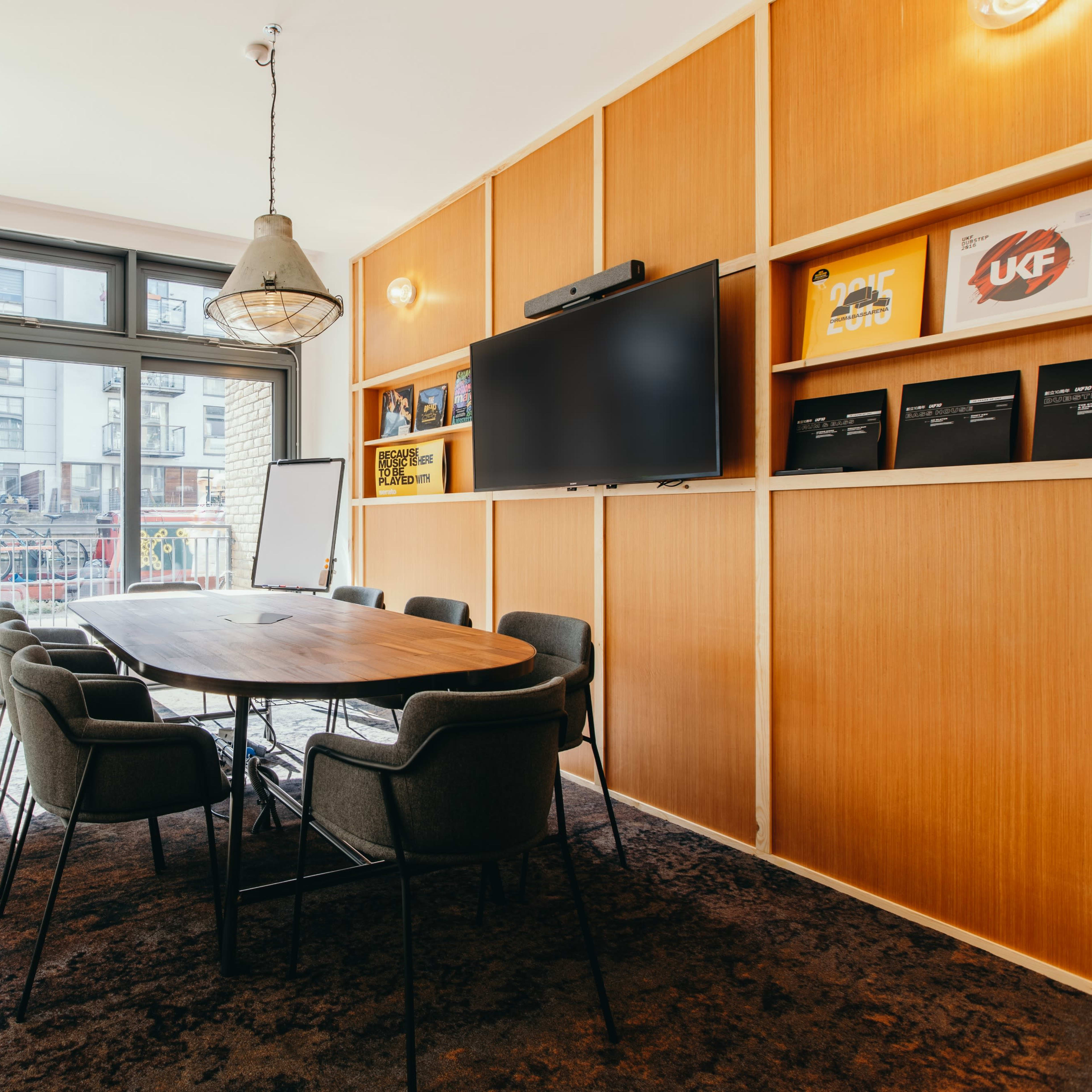 A modern conference room featuring a large wooden table surrounded by six chairs, a wall-mounted television, and shelves displaying various items.