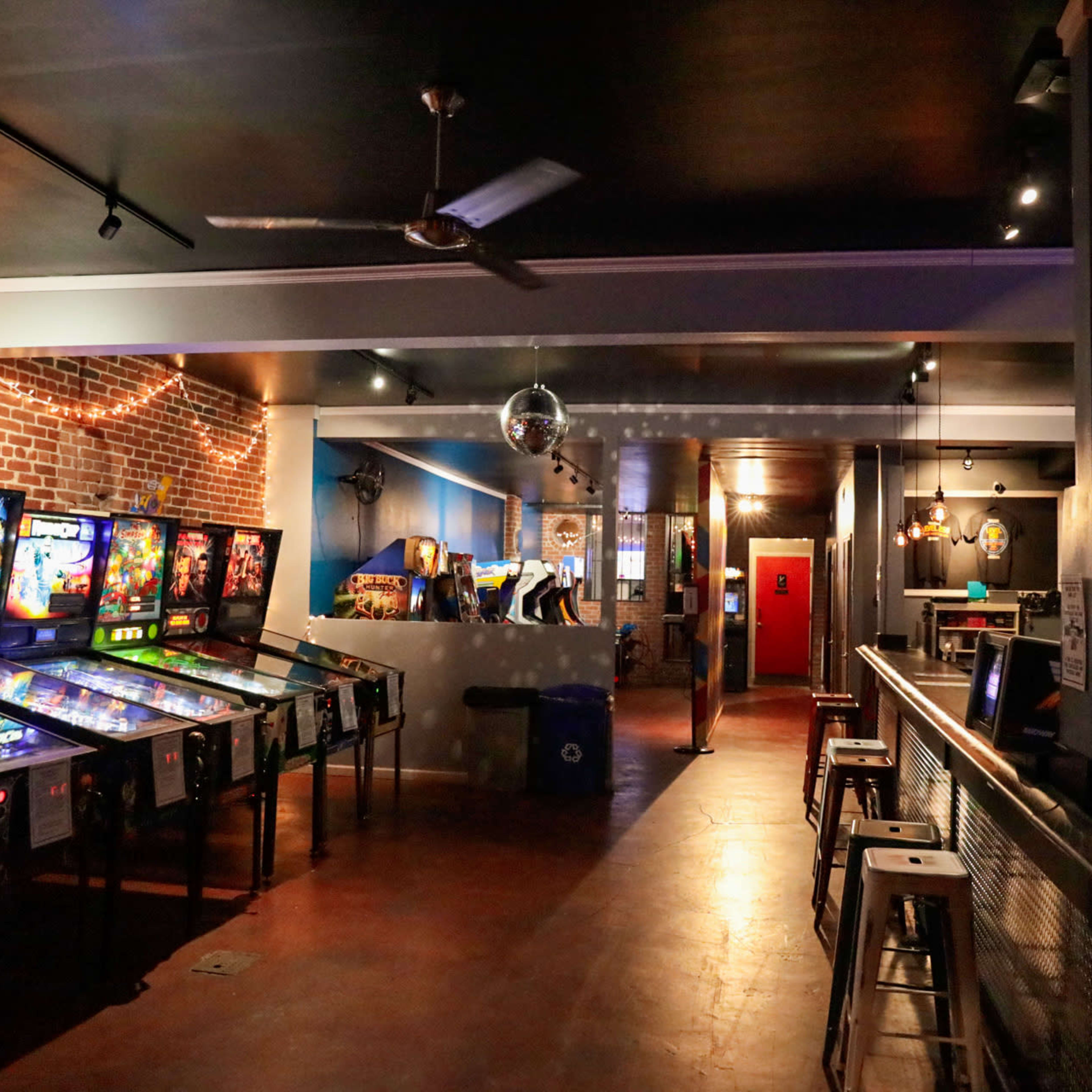 A retro arcade with pinball machines lined up against a brick wall and a bar in the background.