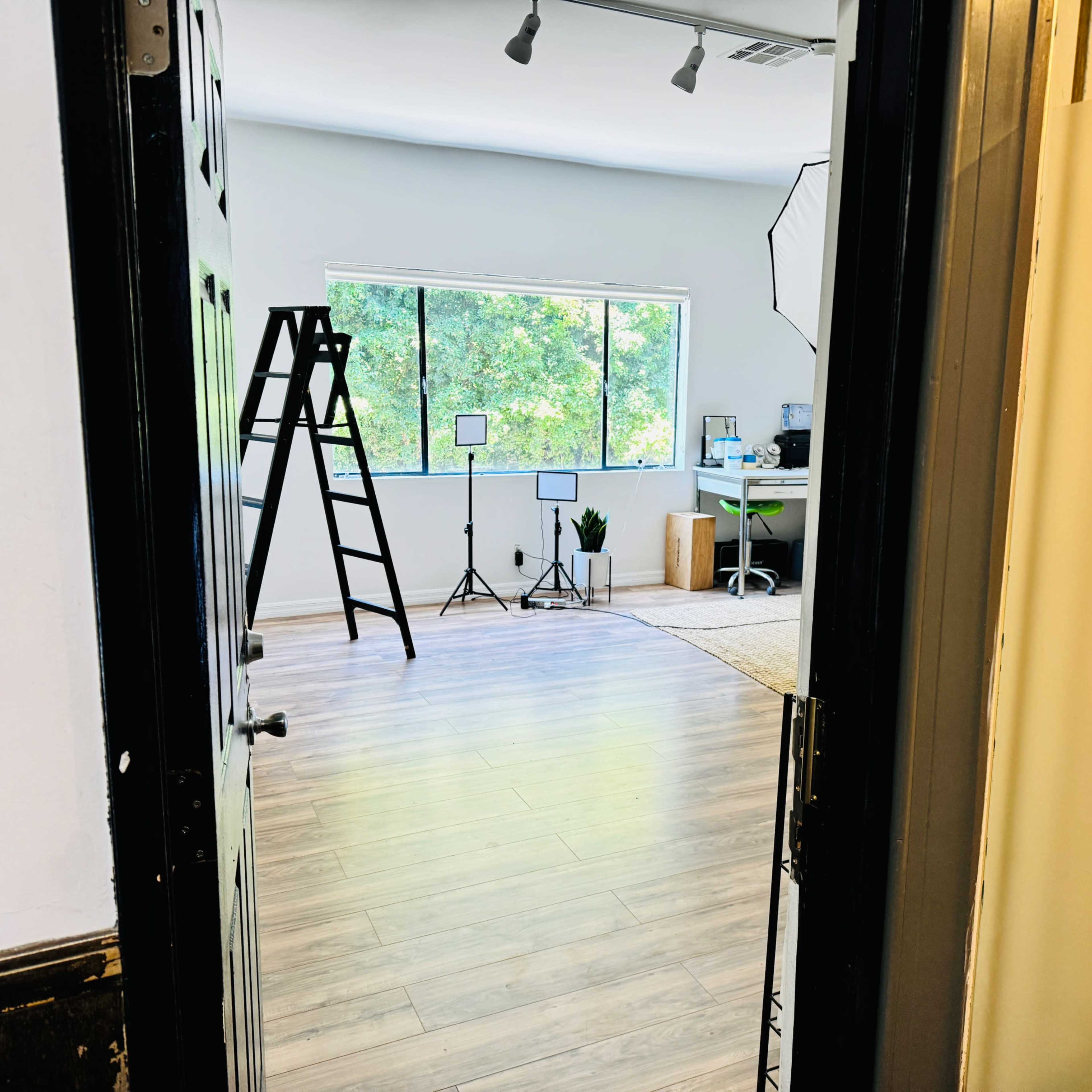 An empty room with large windows, a ladder, a plant, and a desk setup in the corner.