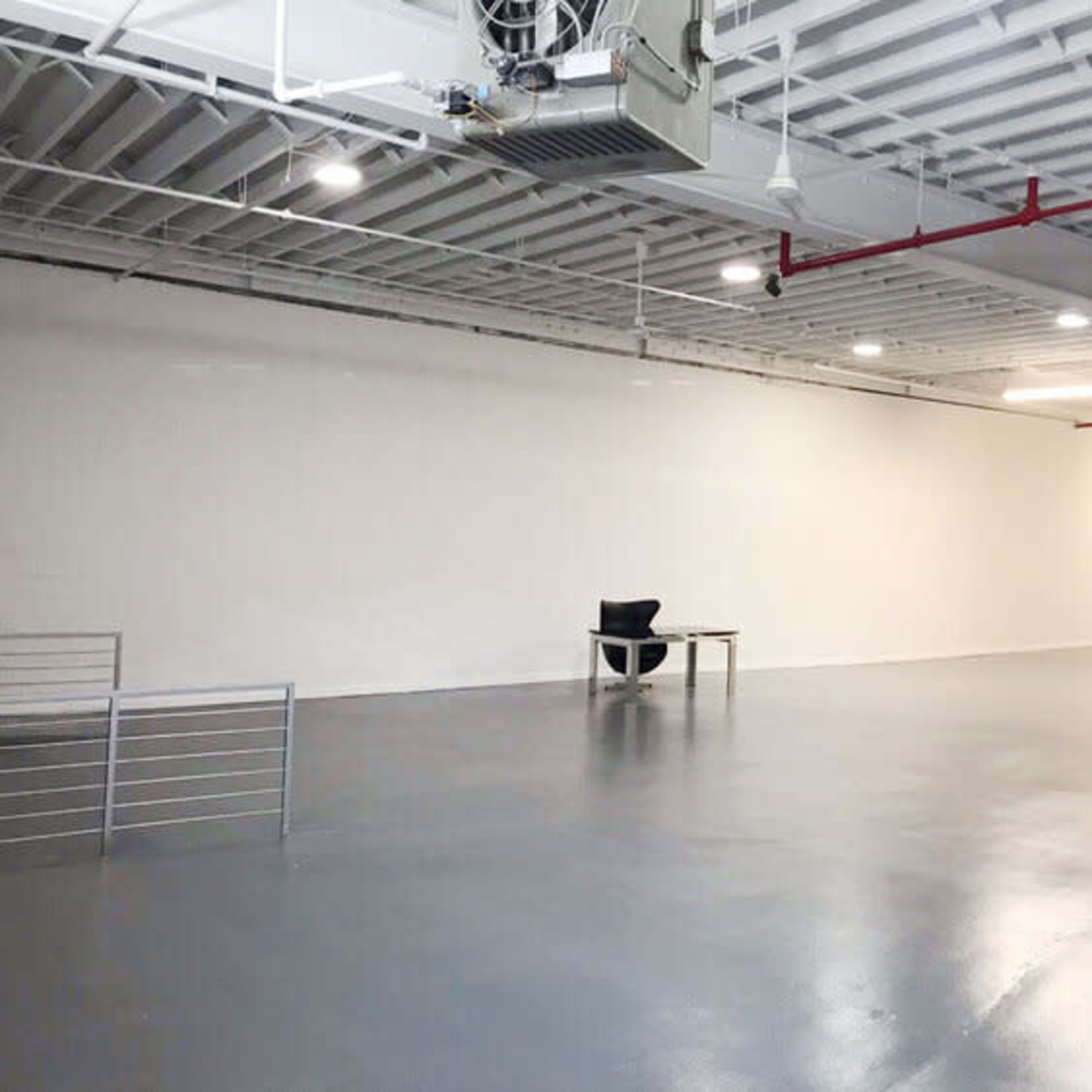 A large, empty room with a polished floor, featuring a single black chair and a table against a plain white wall.