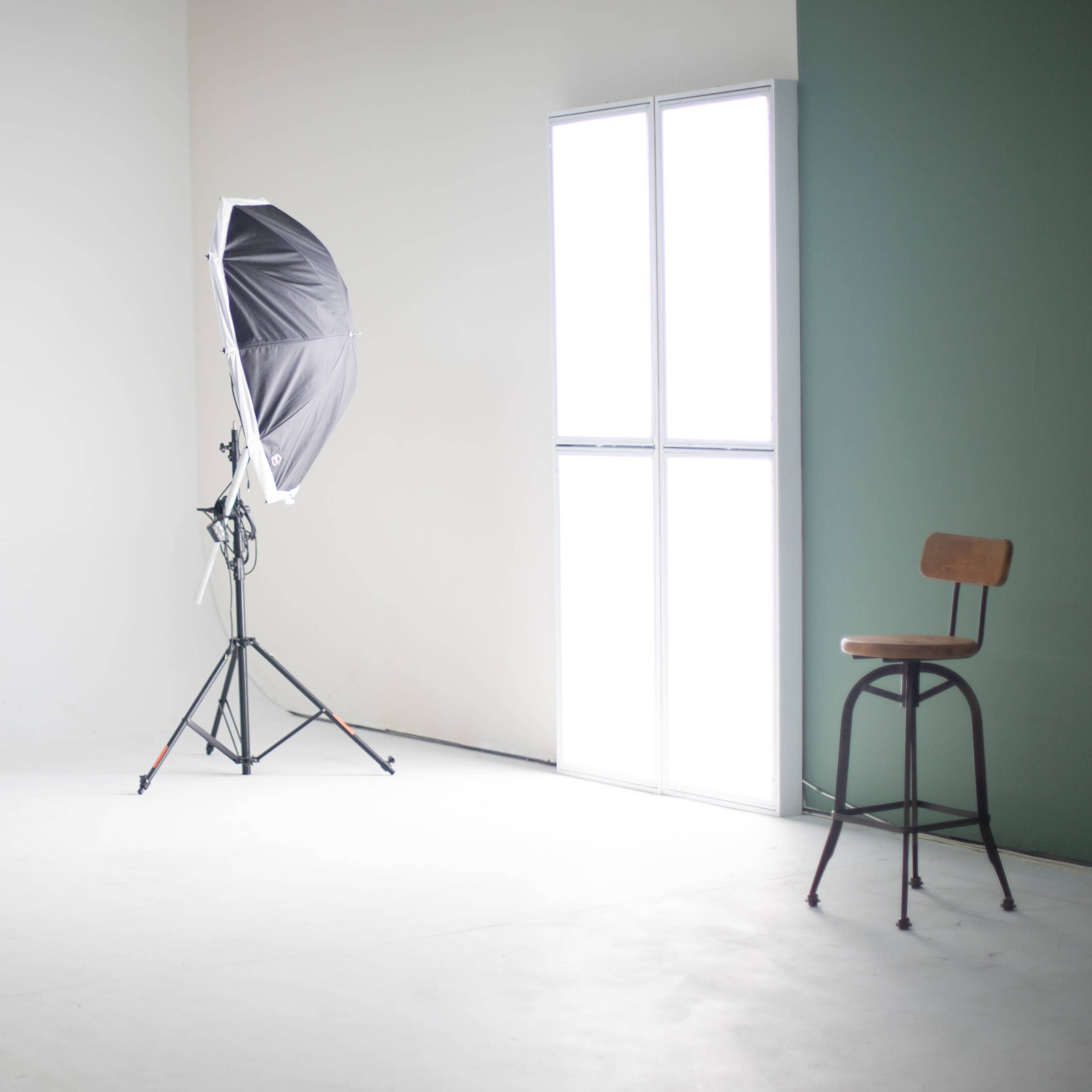 A minimalist studio space with a black softbox light, a tall chair, a low stool, and a large window emitting natural light.
