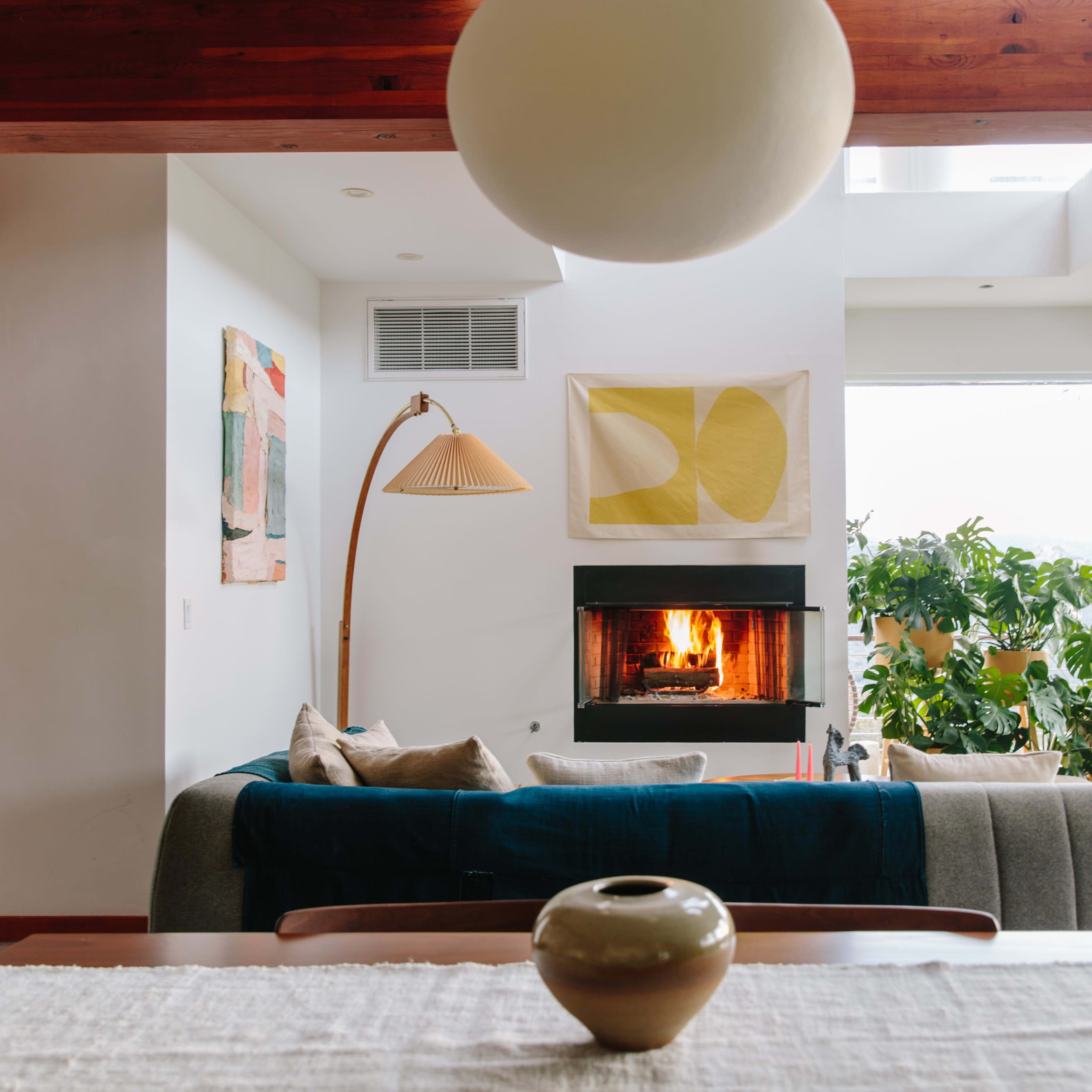 A spacious living room features a gray sofa, a floor lamp, a fireplace with a lit fire, and large windows with a view of greenery.
