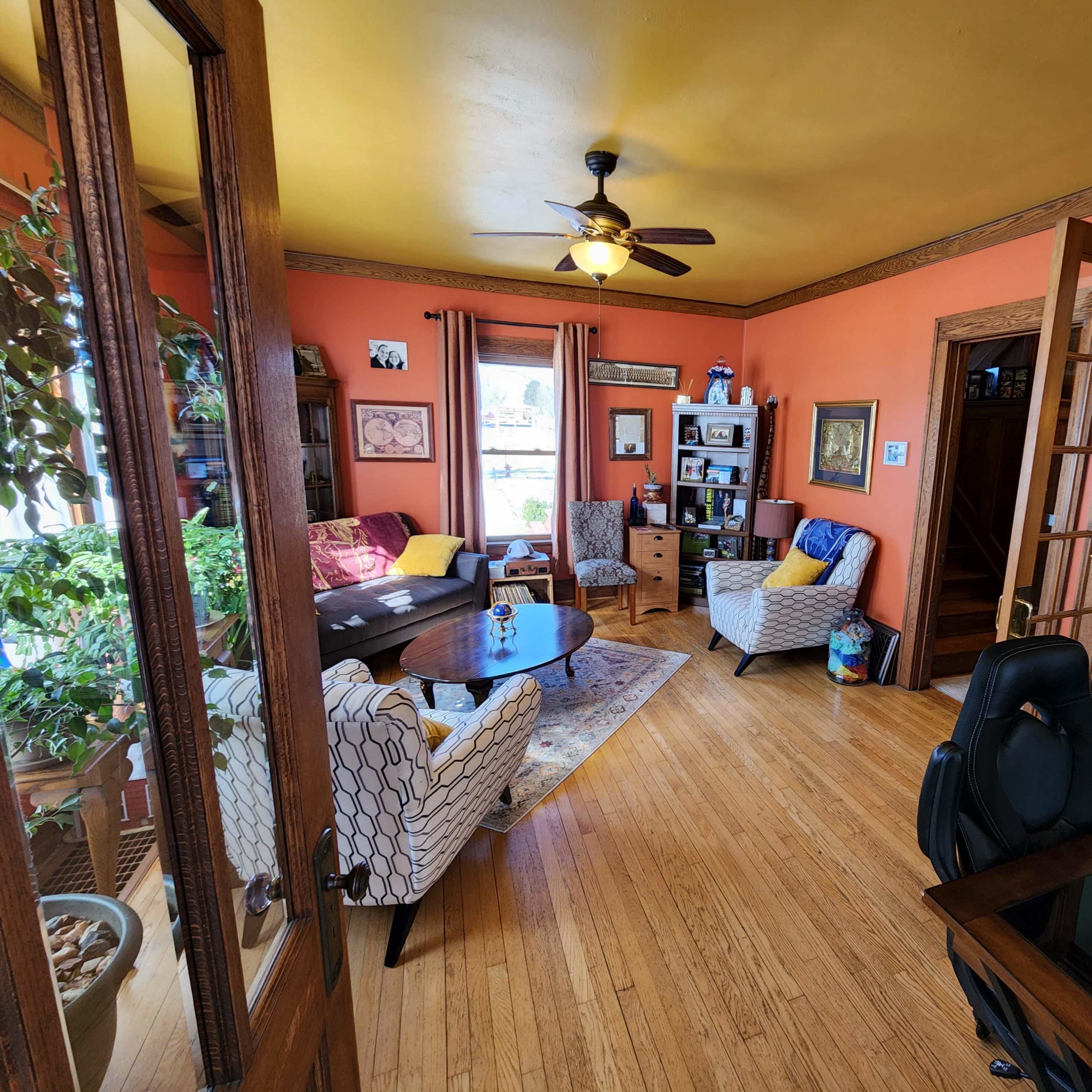A living room with orange walls, hardwood floors, two patterned chairs, a coffee table, a potted plant, and a desk in the foreground.