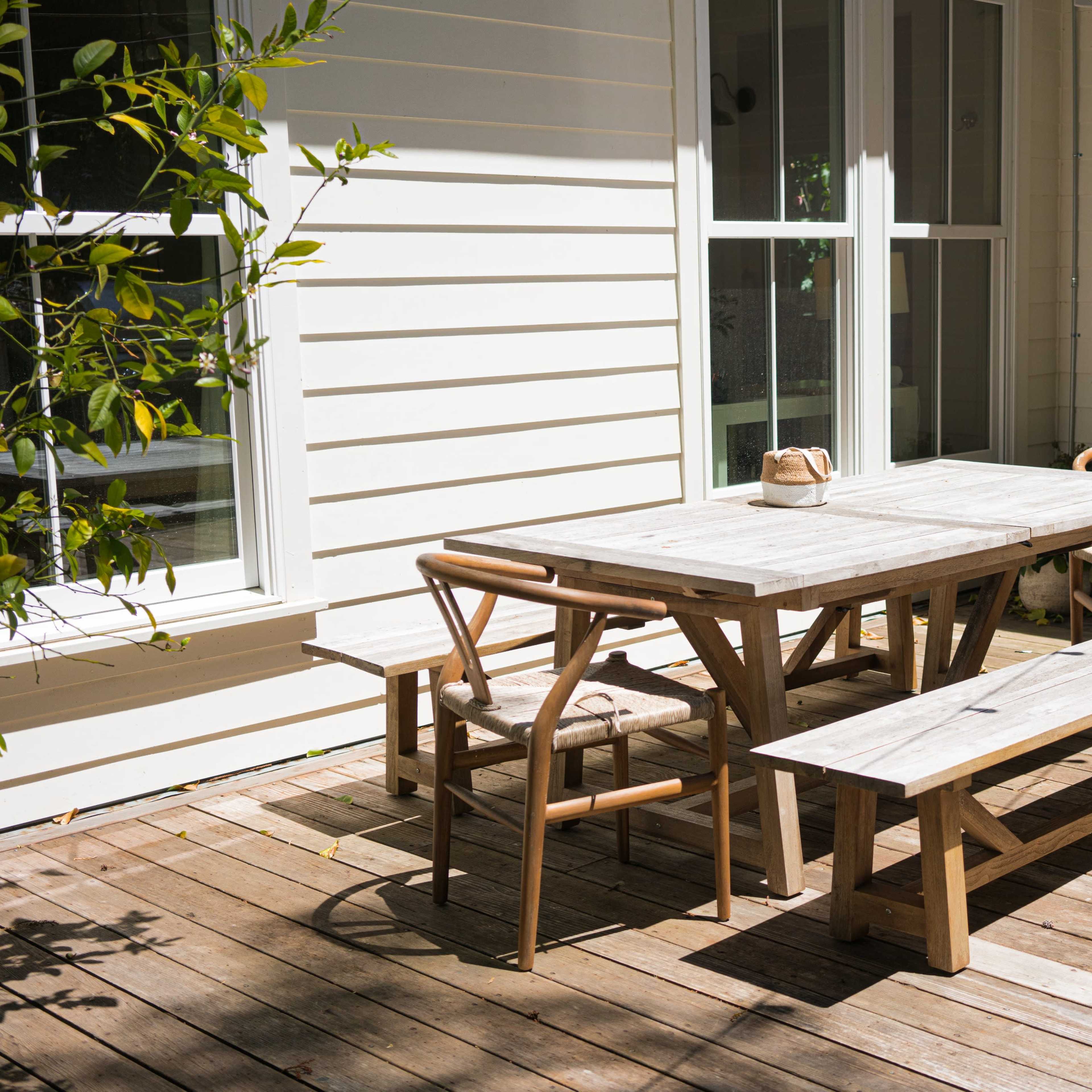 A wooden dining table surrounded by chairs and a bench is positioned on a wooden deck beside a house, with sunlight filtering through nearby plants.