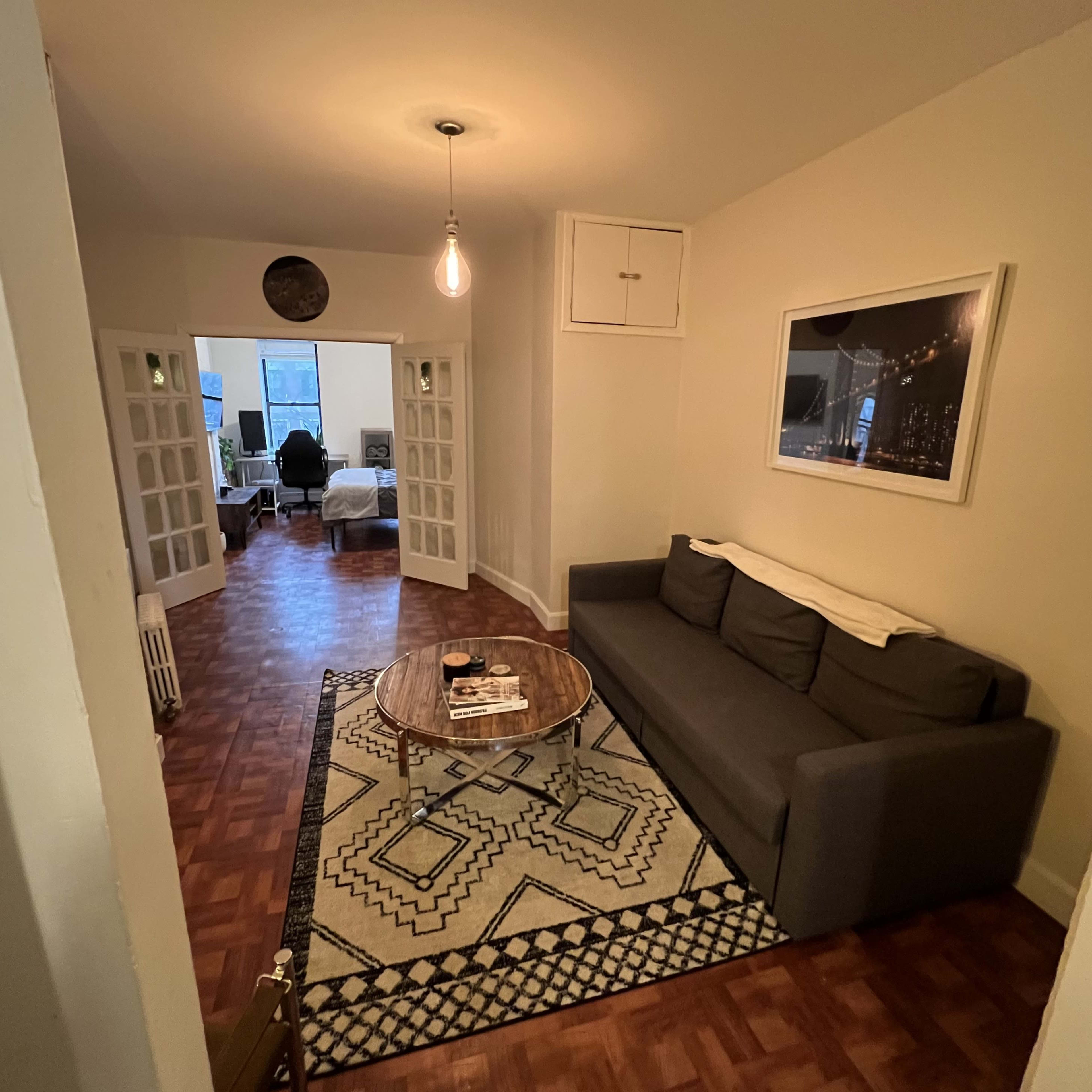 A living room with a gray couch, a round wooden coffee table on a patterned rug, and a glimpse of another room through glass doors.