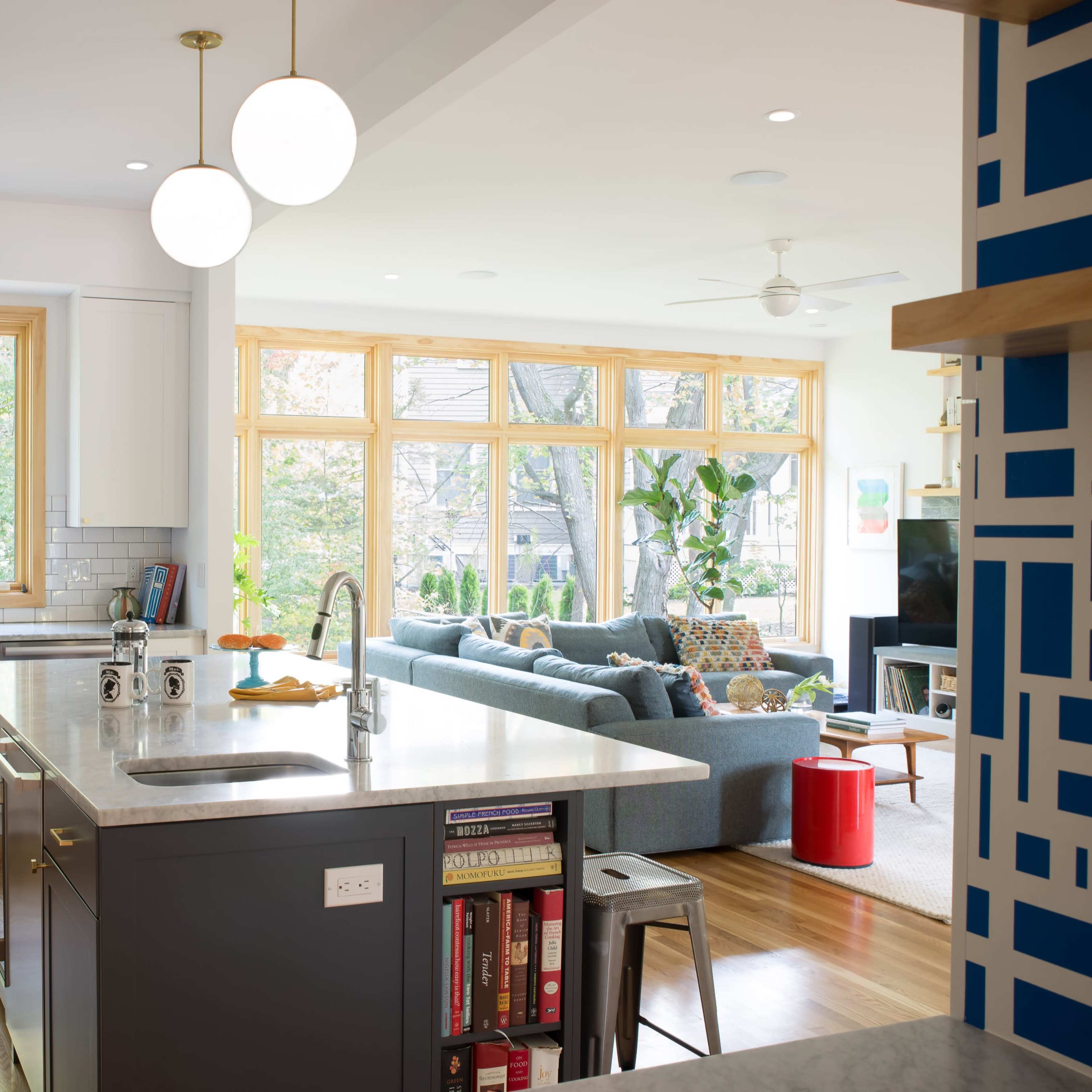 A modern kitchen features a large island, stainless steel appliances, and ample natural light from large windows, flowing into a living area with a sofa and decorative plants.