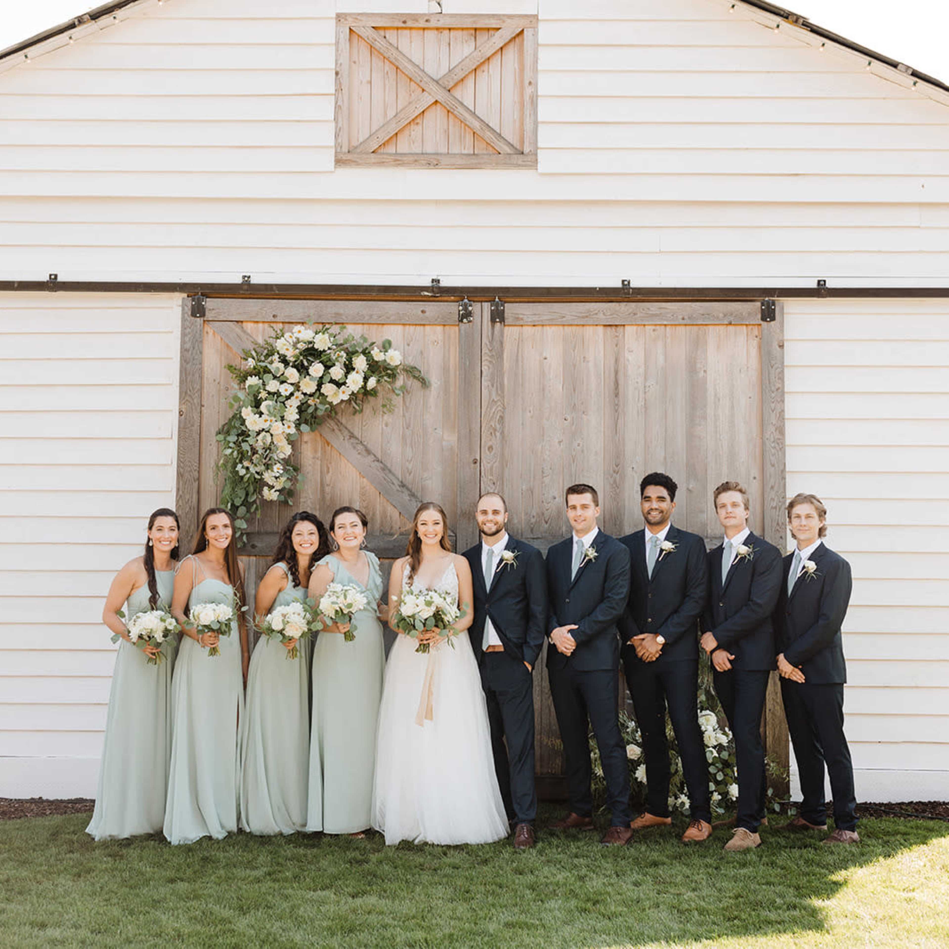 A wedding party poses together in front of a white barn with wooden doors adorned with floral decorations.