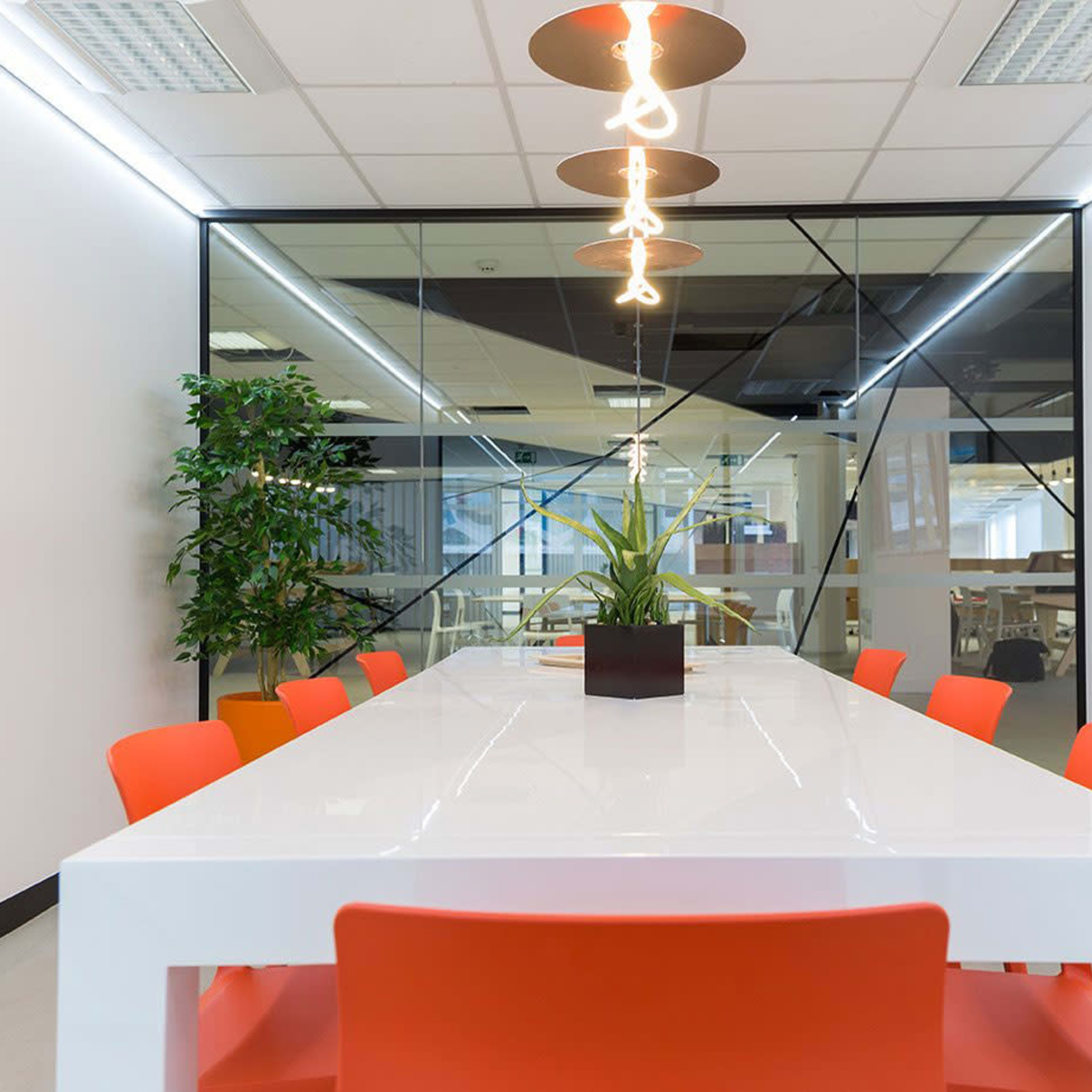 A modern conference room features a long white table surrounded by orange chairs, with a plant centerpiece and a large glass wall in the background.