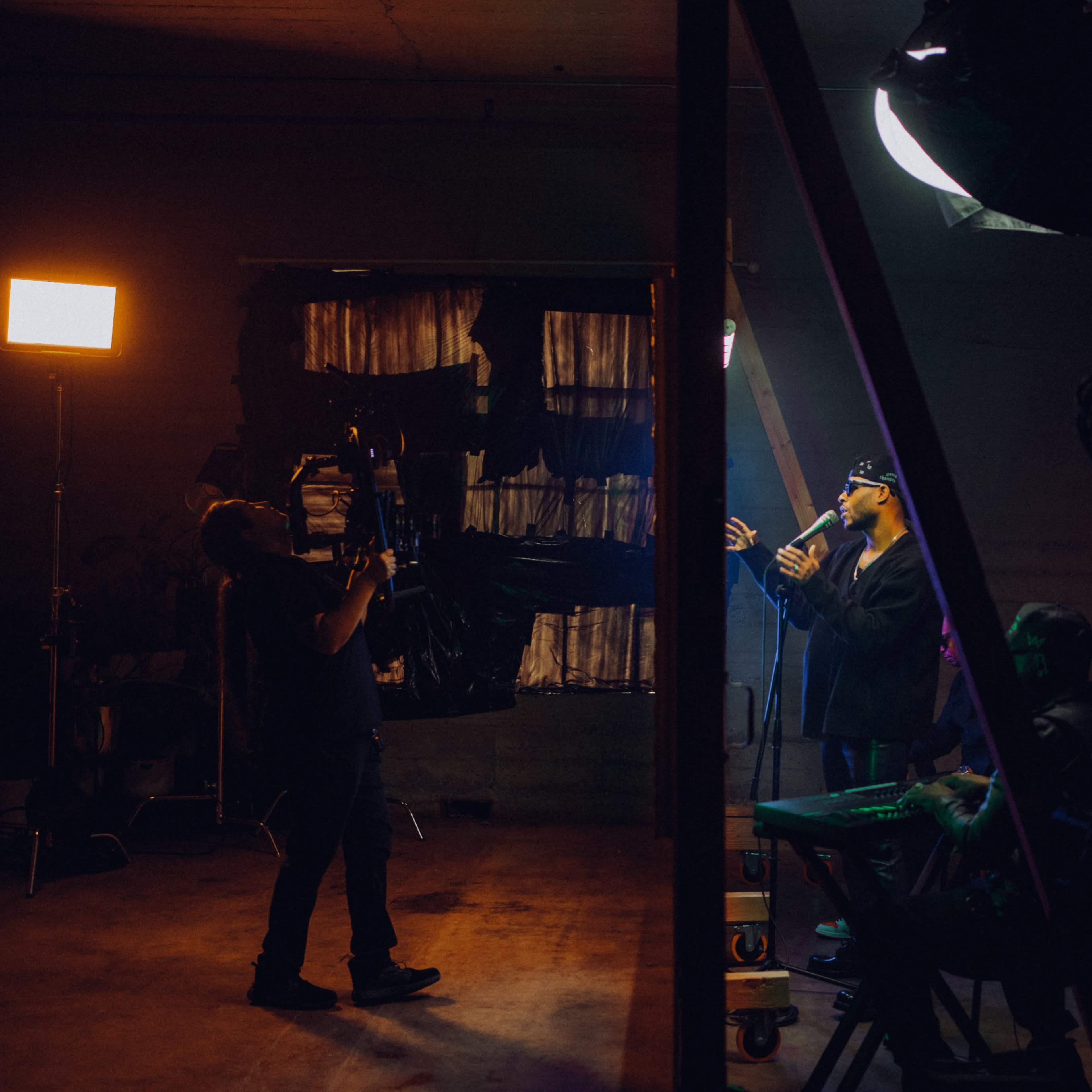 A musician performs on stage while a cameraman films the scene in a dimly lit studio.