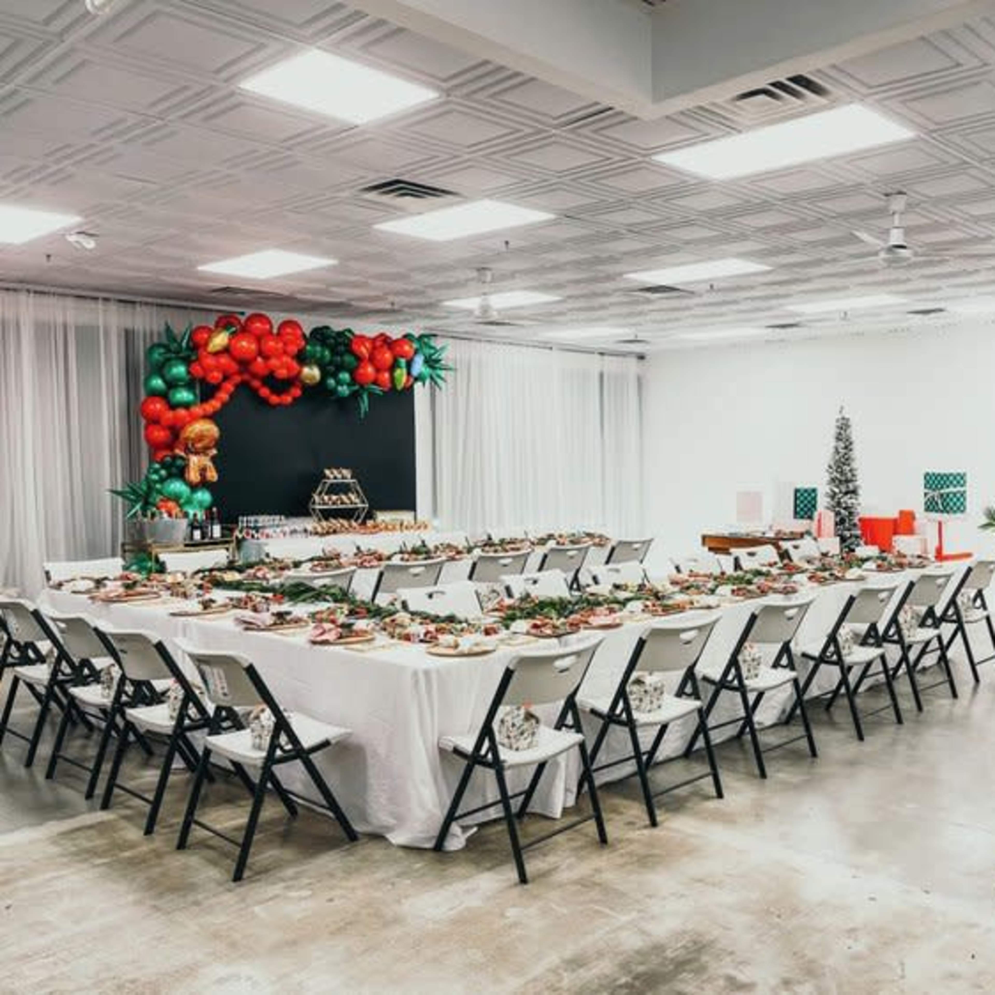 A large dining area is set up with multiple tables covered in white cloth and arranged for a festive gathering, featuring holiday decorations and a buffet setup.