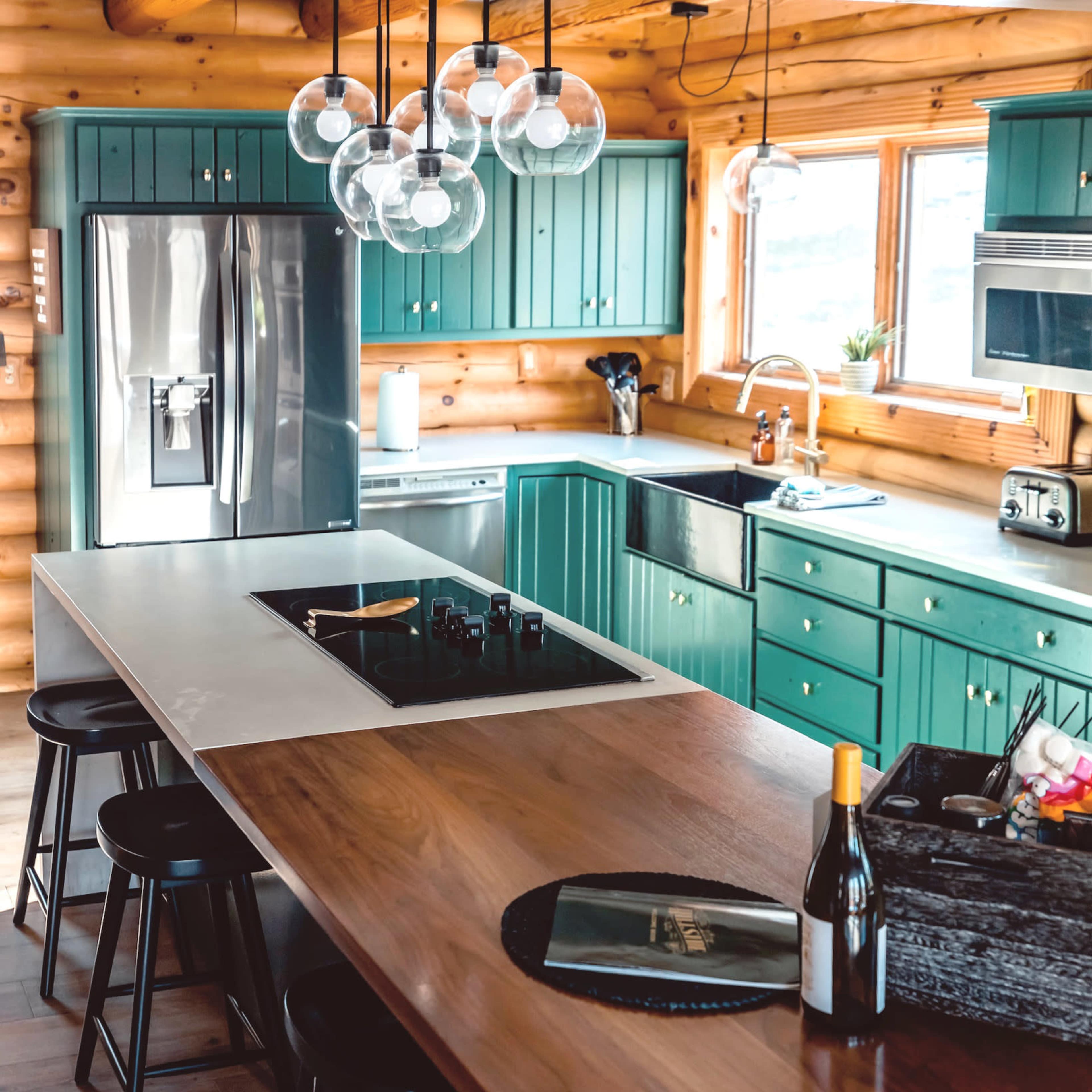 A modern kitchen features teal cabinetry, a central island with a wooden countertop, and pendant lighting above bar stools.