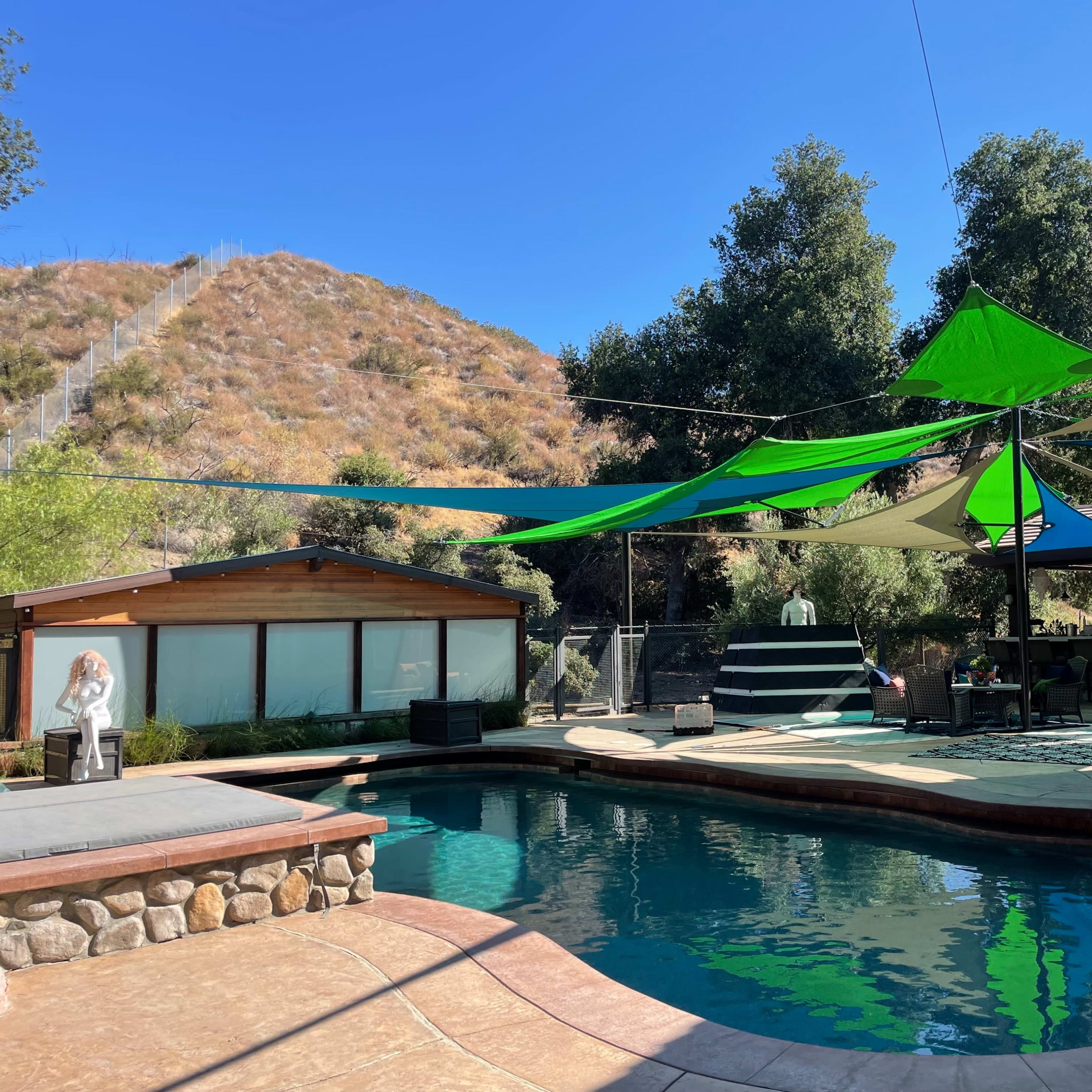 A swimming pool area features colorful fabric shades overhead, surrounded by green trees and a hillside in the background.