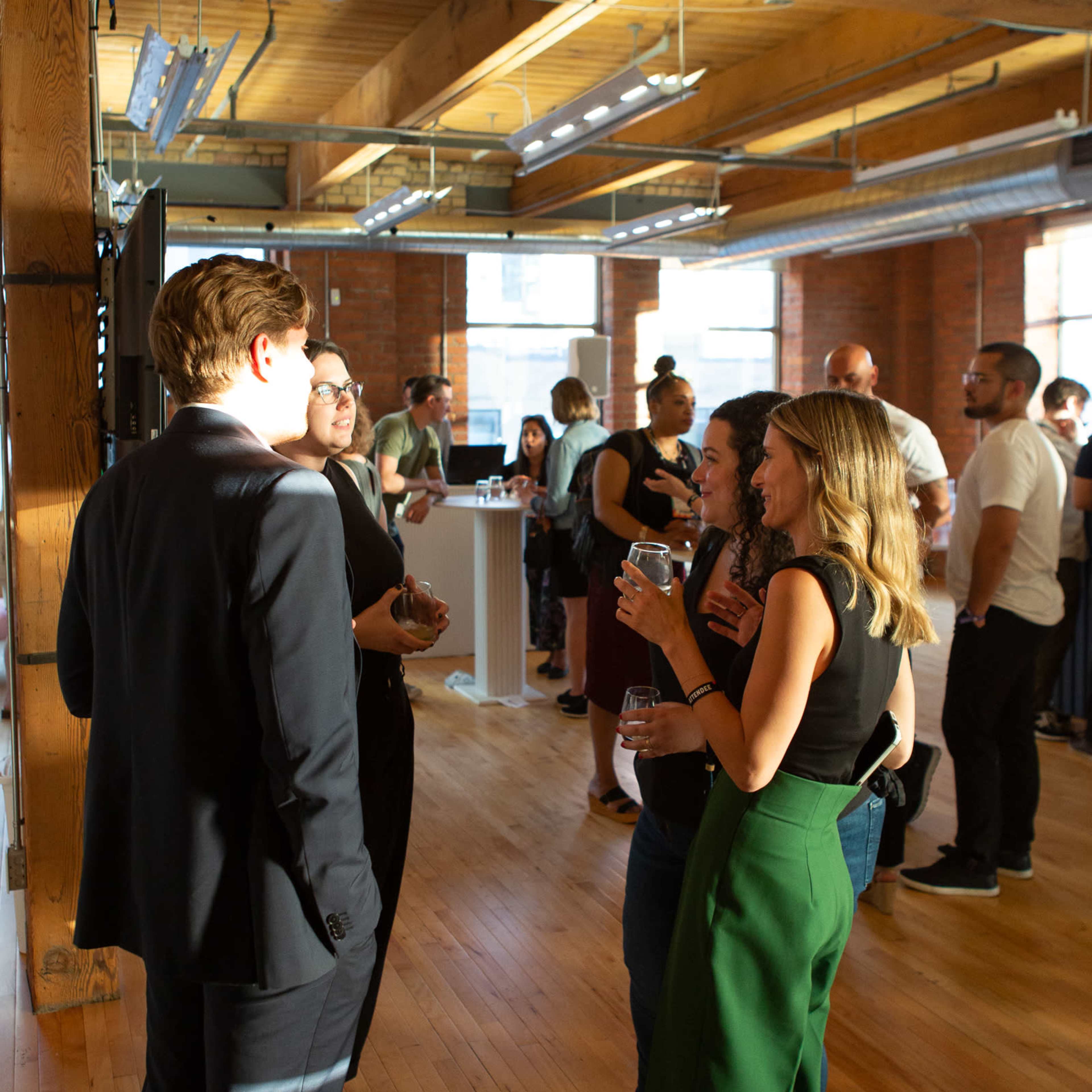 A group of people engage in conversation at a social event held in a spacious, well-lit room with wooden floors and exposed beams.