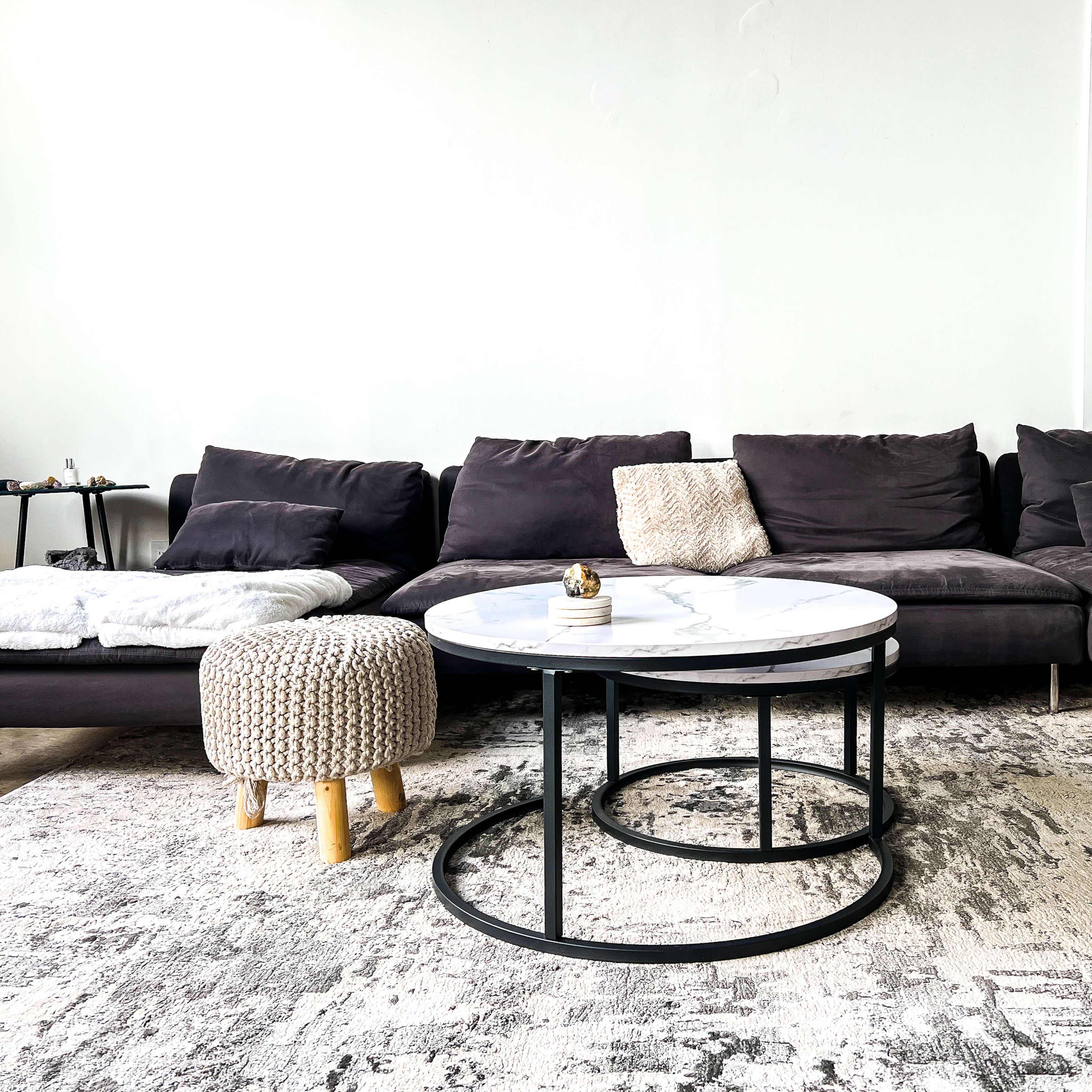 A modern living room features a dark gray sectional sofa, a round marble coffee table with metal legs, and a light-colored knitted pouf on a large area rug.