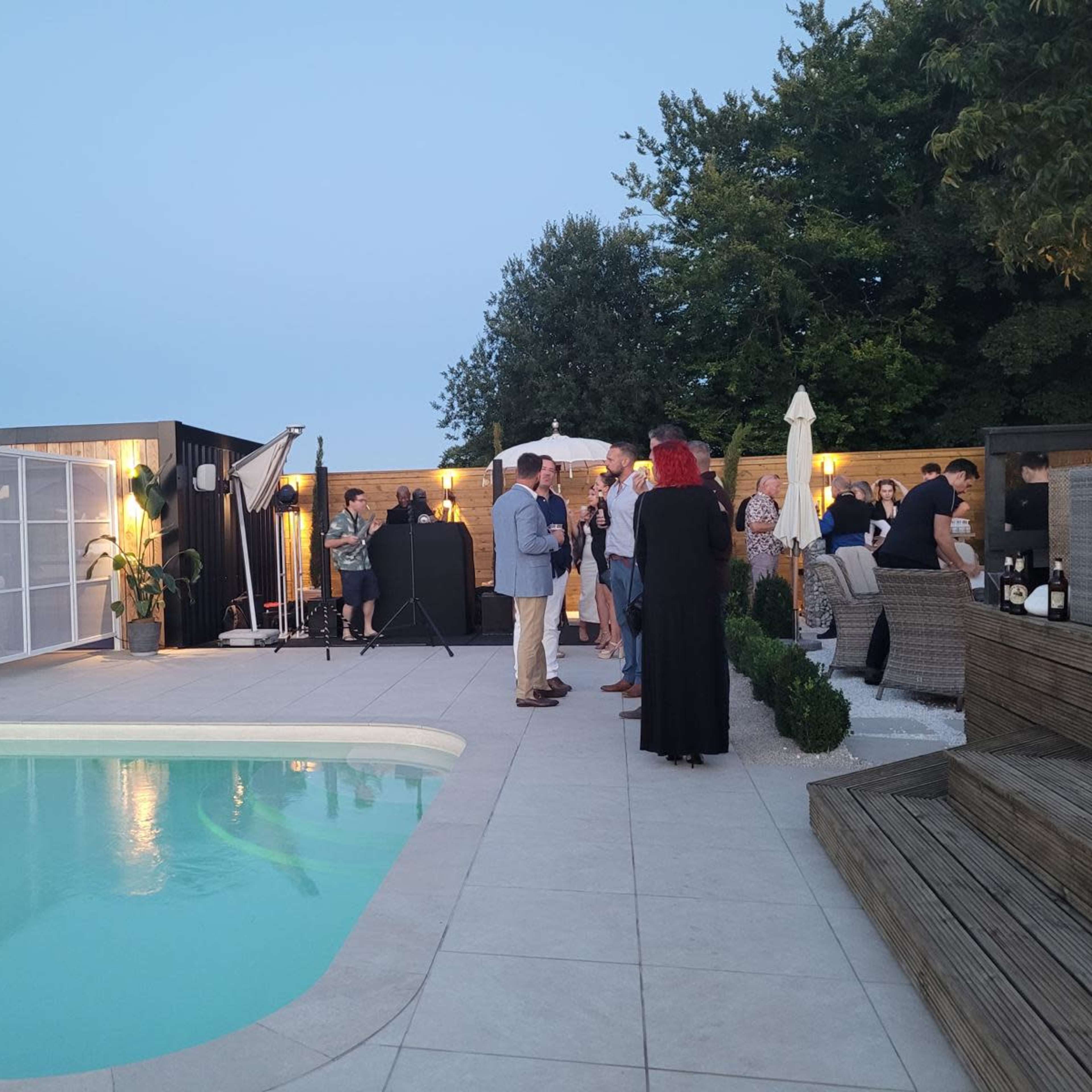 A group of people socialize around a swimming pool in a backyard setting during the evening.