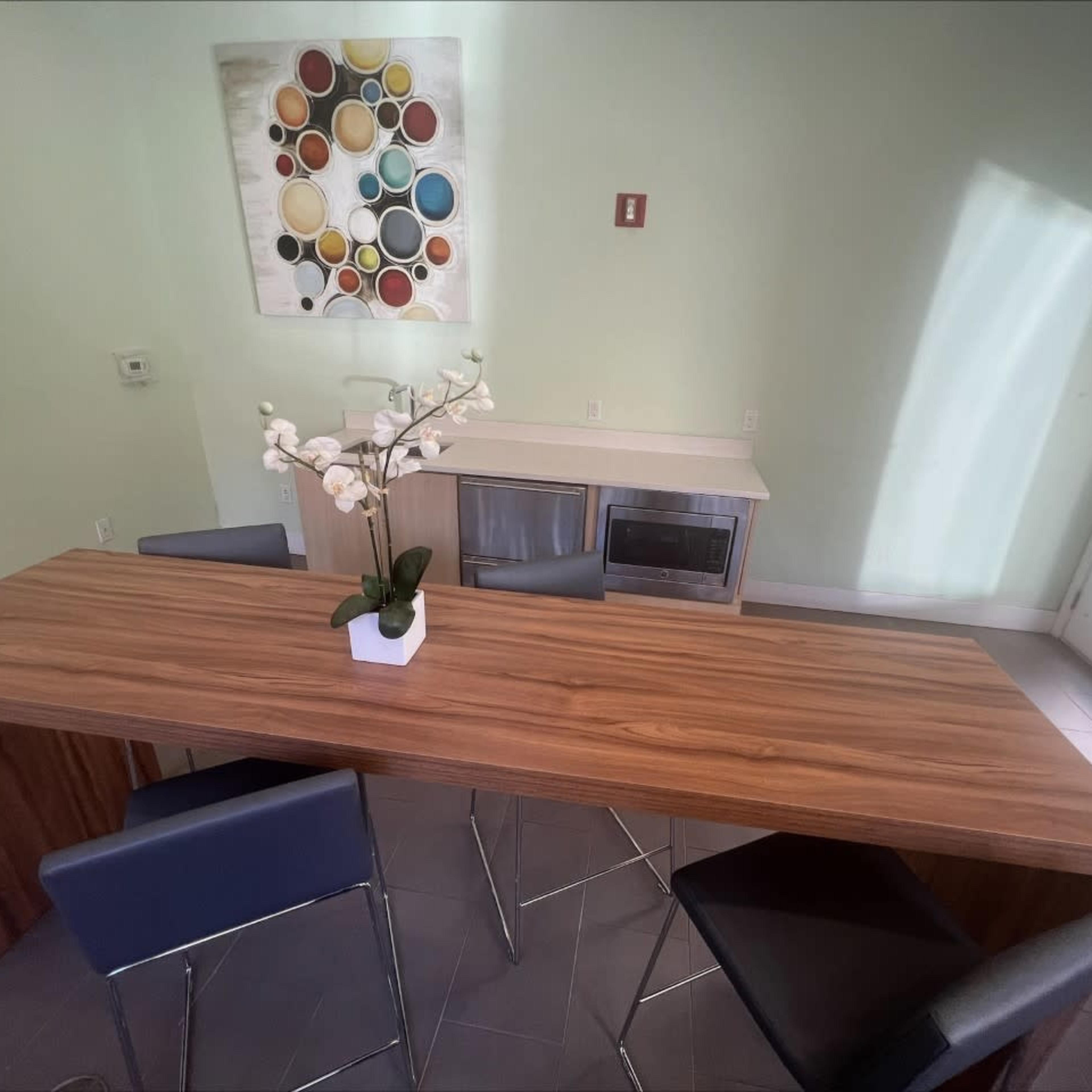 A wooden dining table with black chairs is positioned in a light-colored room, featuring a cabinet and a decorative wall piece above.